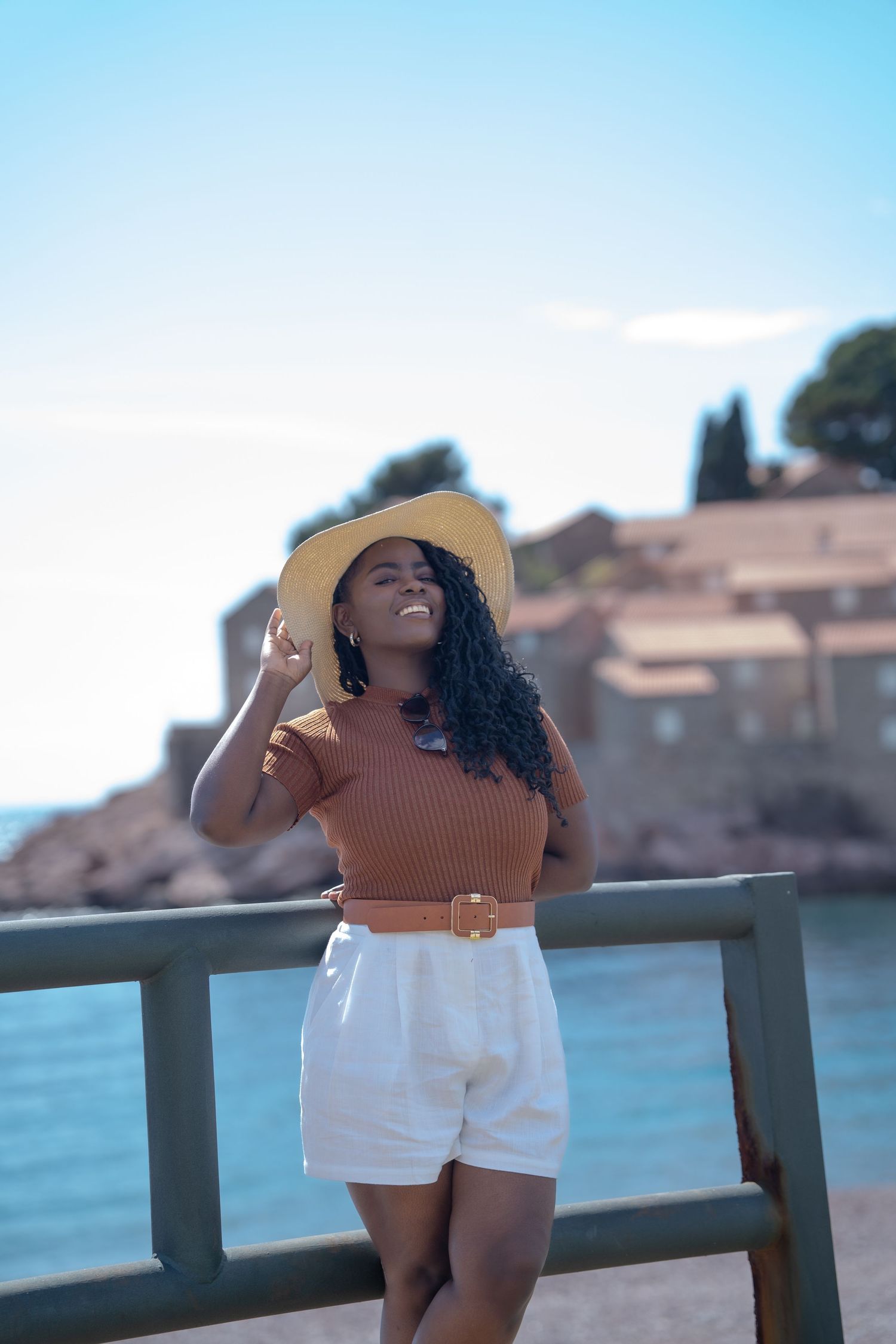 Person in a brown top and white shorts enjoying a sunny day by the waterfront, wearing a straw hat.