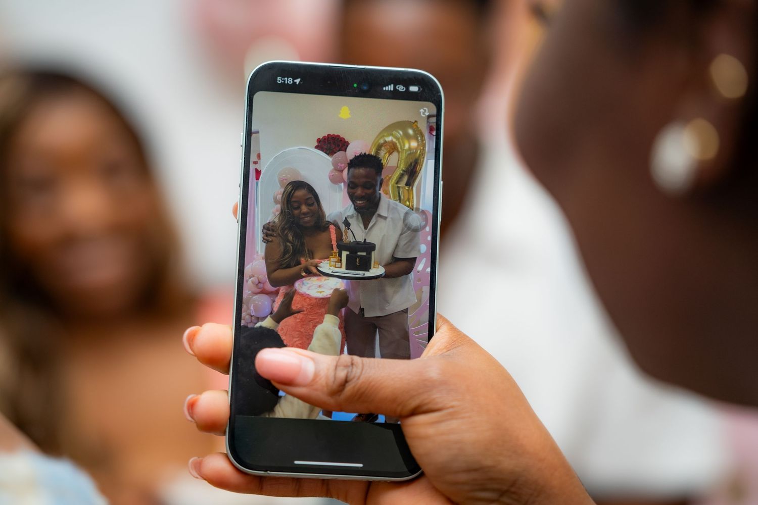 Close-up view of hands holding a smartphone displaying people looking at a tablet screen during a gathering.
