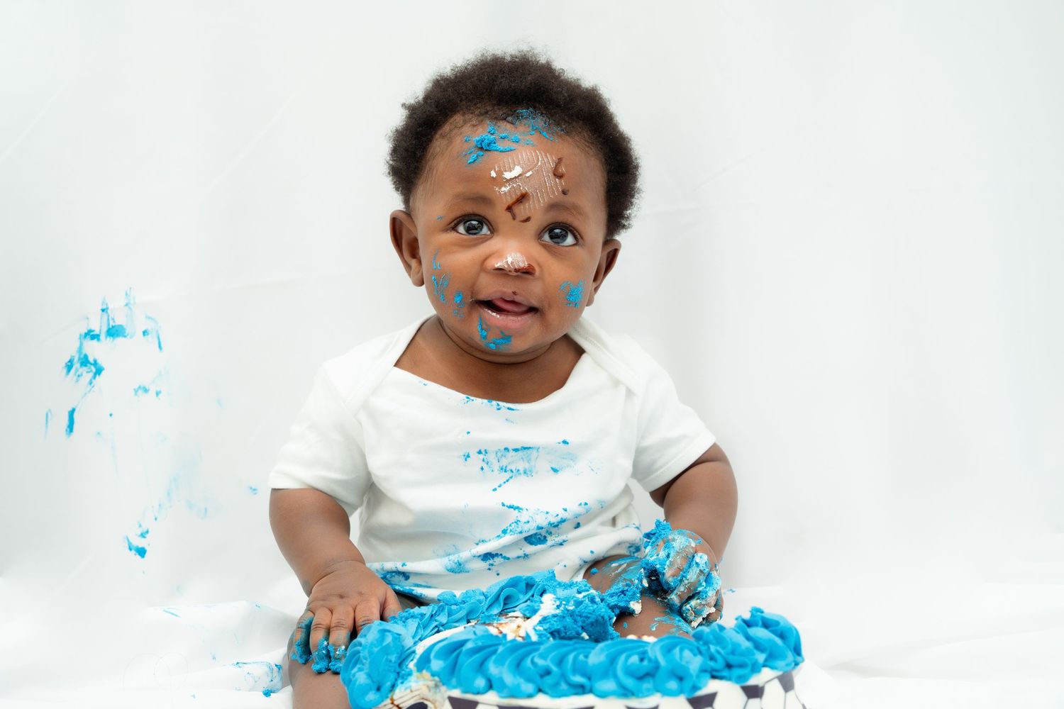 Small child wearing white shirt sits on floor with bright blue paint smeared on face and clothes against white backdrop.