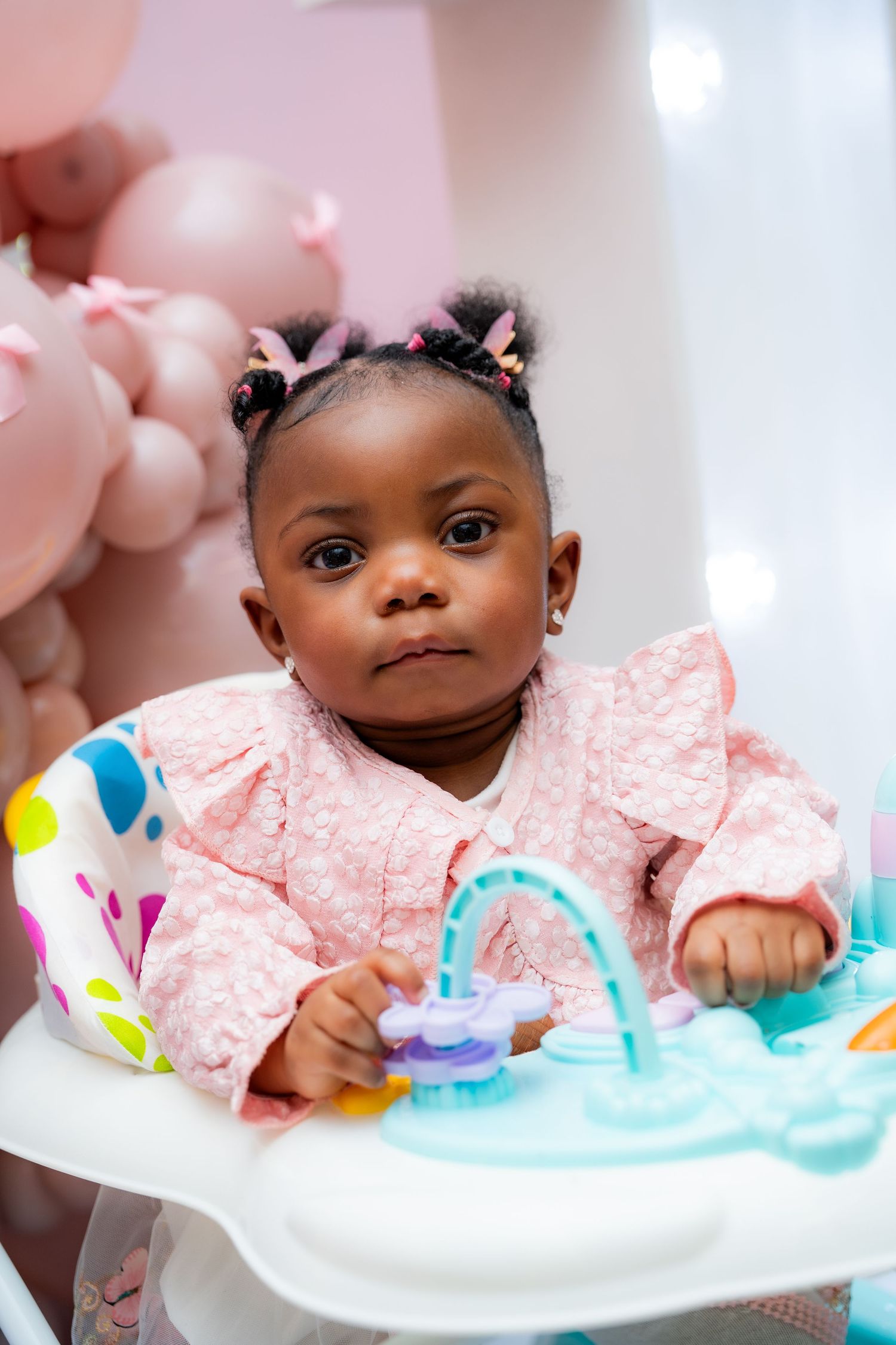 Young child in pink dress sits in activity center with colorful toys and decorations in background.
