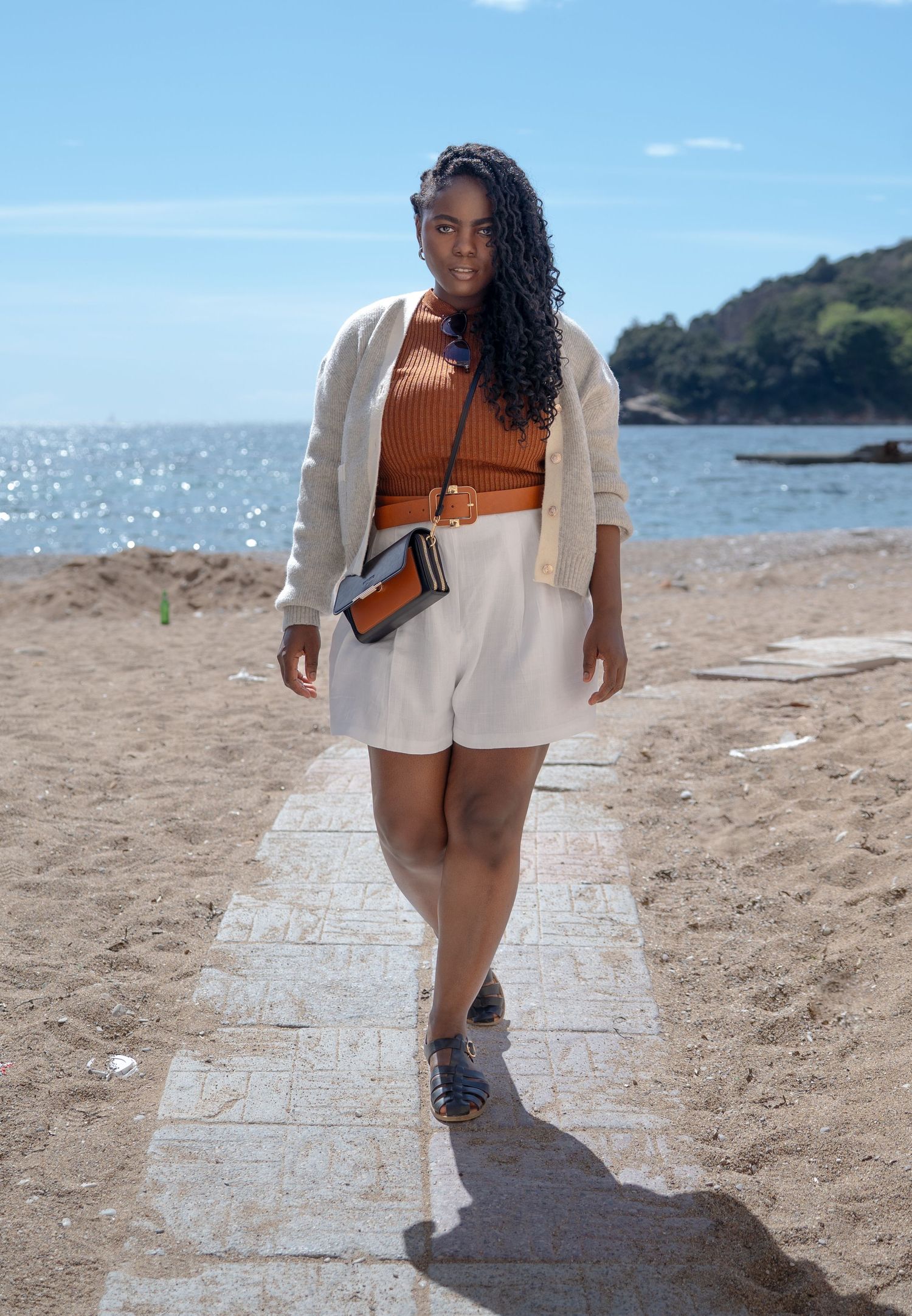 Person in casual summer outfit with cardigan and shorts walks along beach path on sunny day near coastal waters.