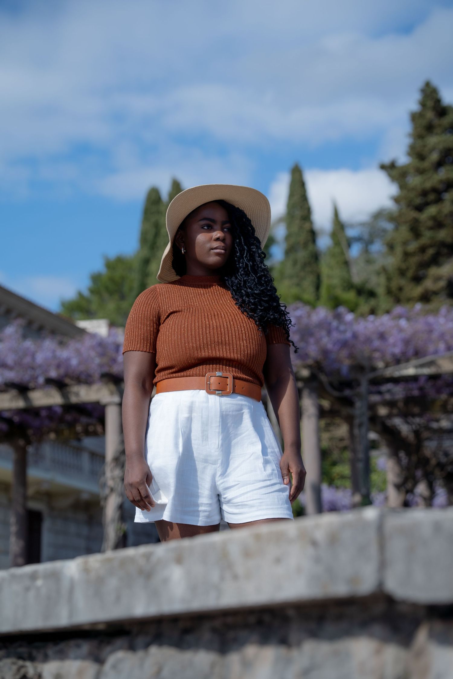 Spring fashion portrait on stone wall with blooming wisteria and cypress trees against blue sky in background.