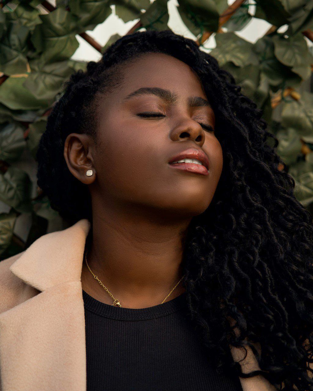 Woman with natural curly hair and beige coat poses against green foliage background in warm sunlight.