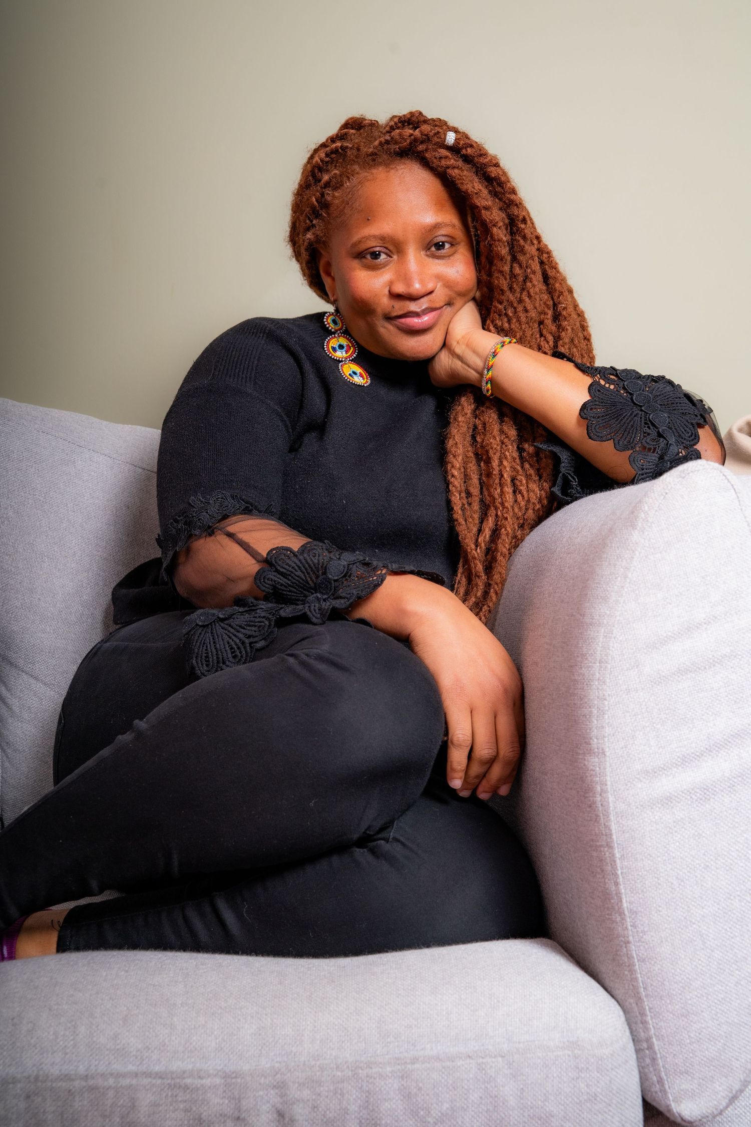 Relaxed person with long auburn locs wearing black sweater lounges comfortably on gray couch.