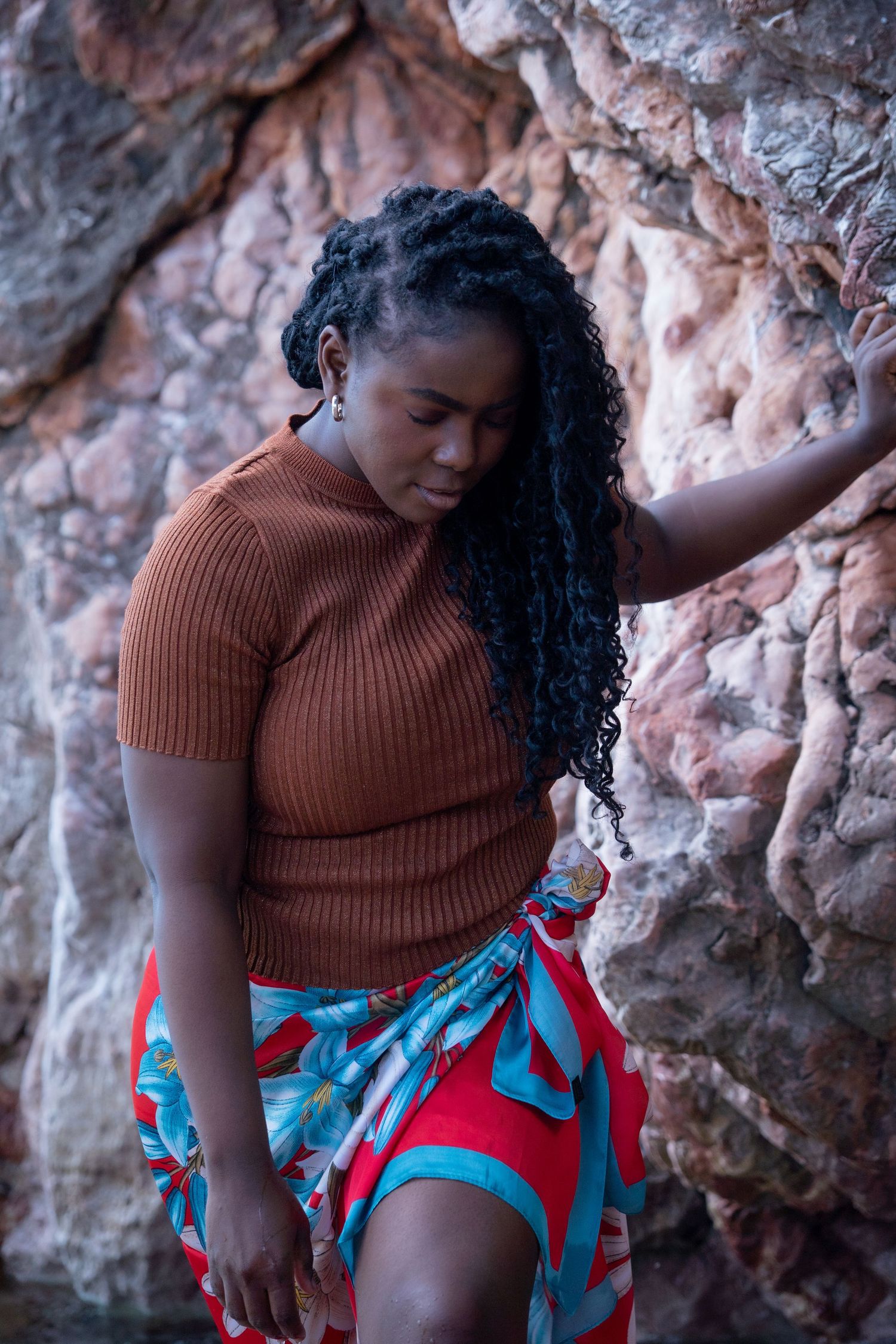 A person in a brown ribbed sweater and patterned skirt stands against a textured rock wall in natural lighting.