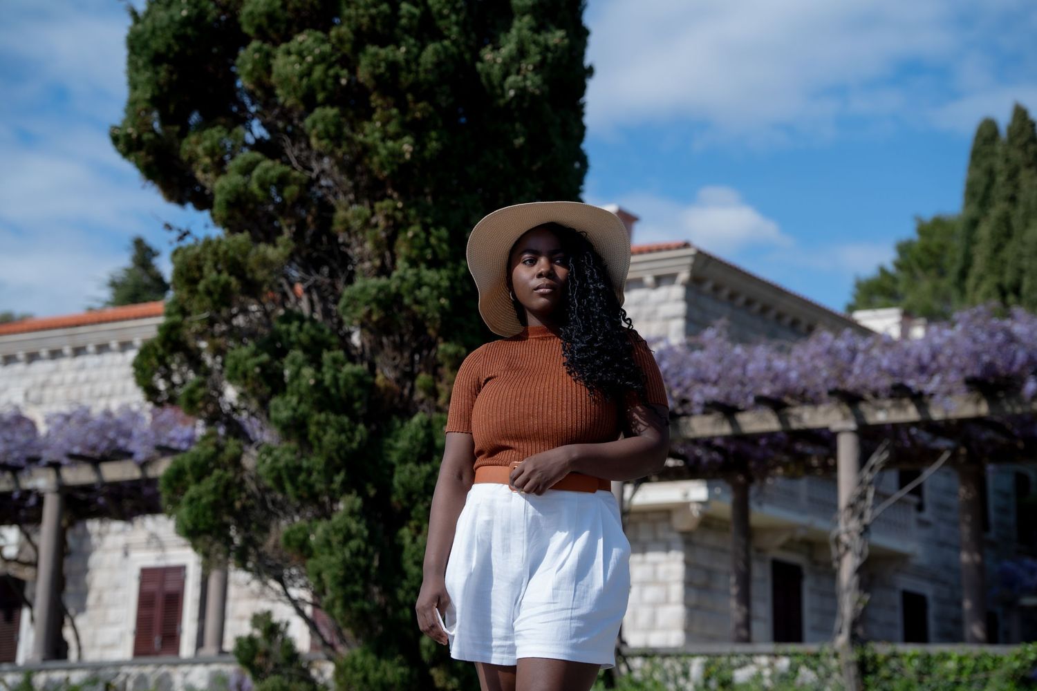Person in a sunhat, rust-colored sweater and white skirt poses in front of a garden with blooming wisteria and buildings.