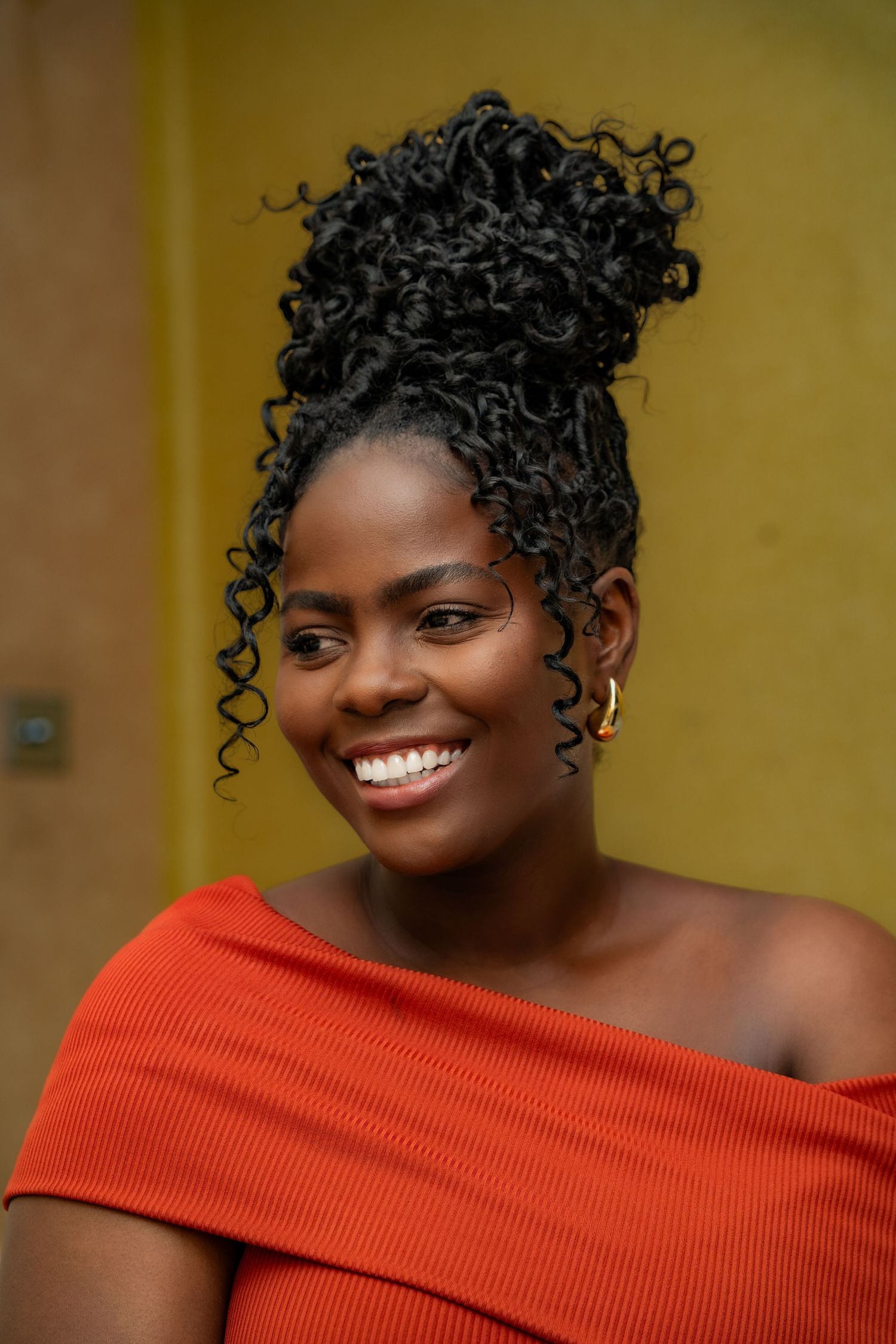 Beautiful natural curly updo hairstyle with cascading tendrils, wearing an elegant off-shoulder orange ribbed top.