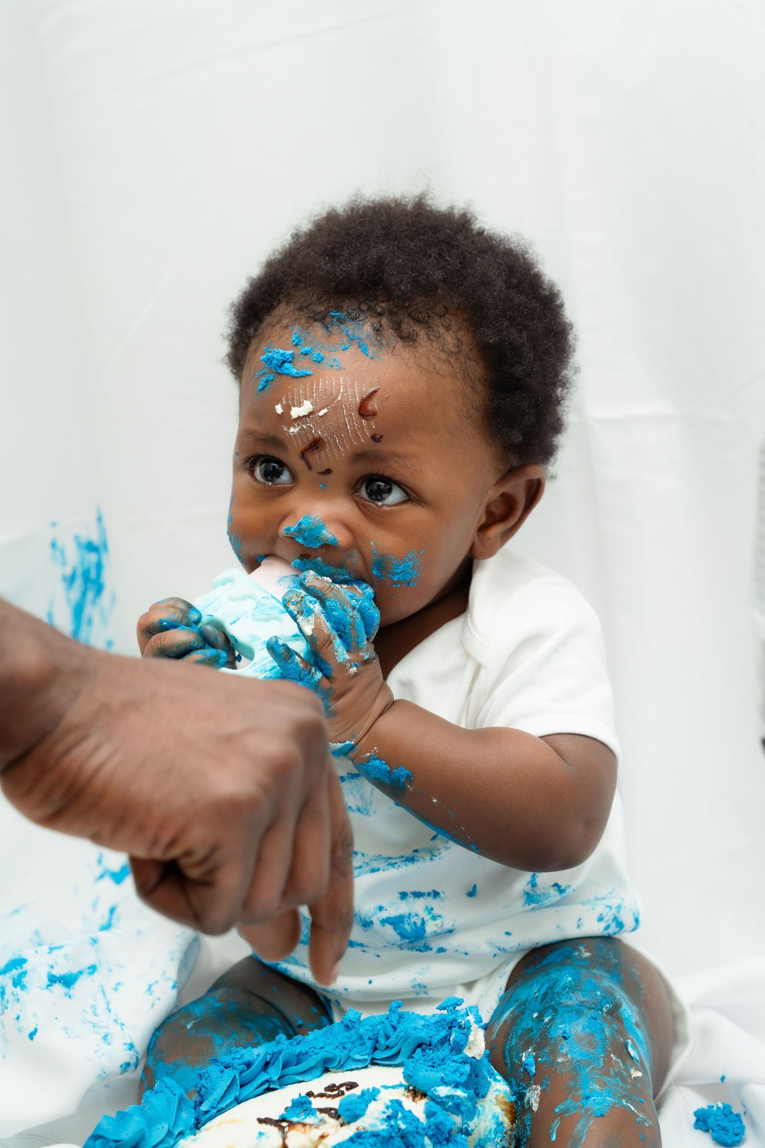 Baby with blue frosting smeared on face and clothing enjoys first birthday cake smash photo session.