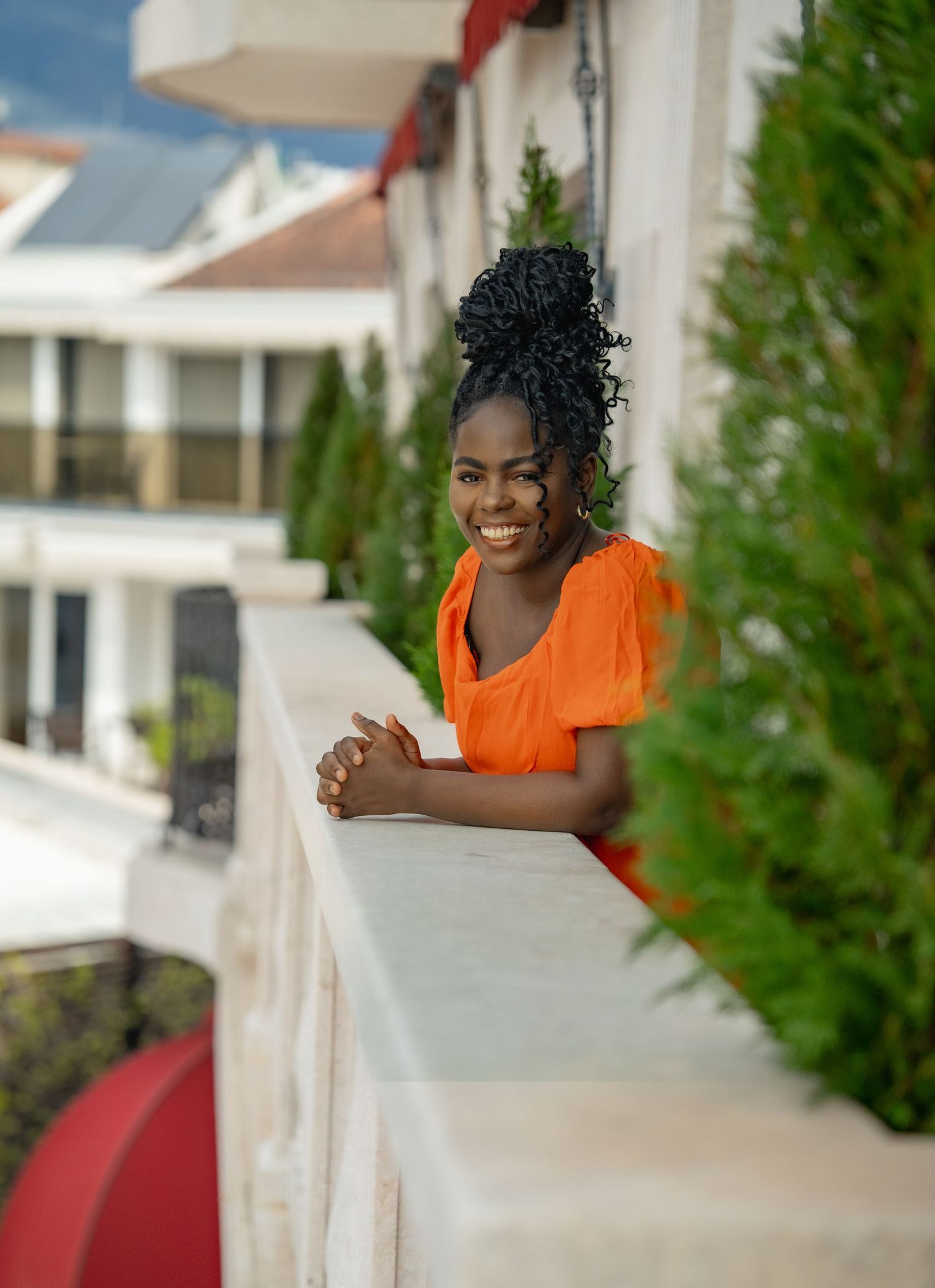 Someone in a bright orange top leans against a white railing with a sincere smile, with evergreen trees in the background.