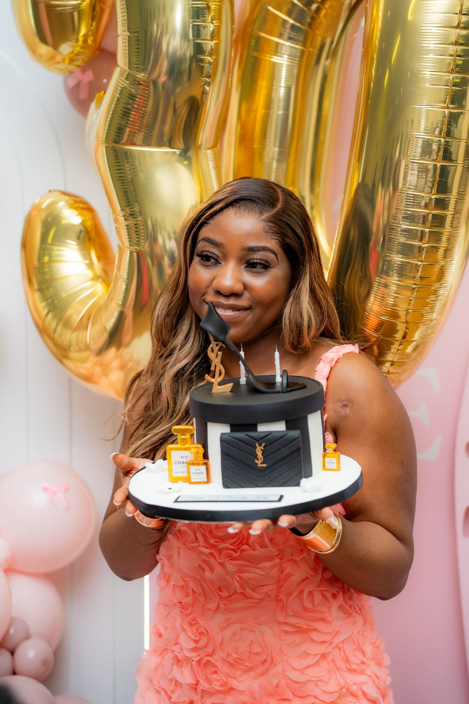 Someone holds a black designer handbag-themed birthday cake with gold balloons in the background.