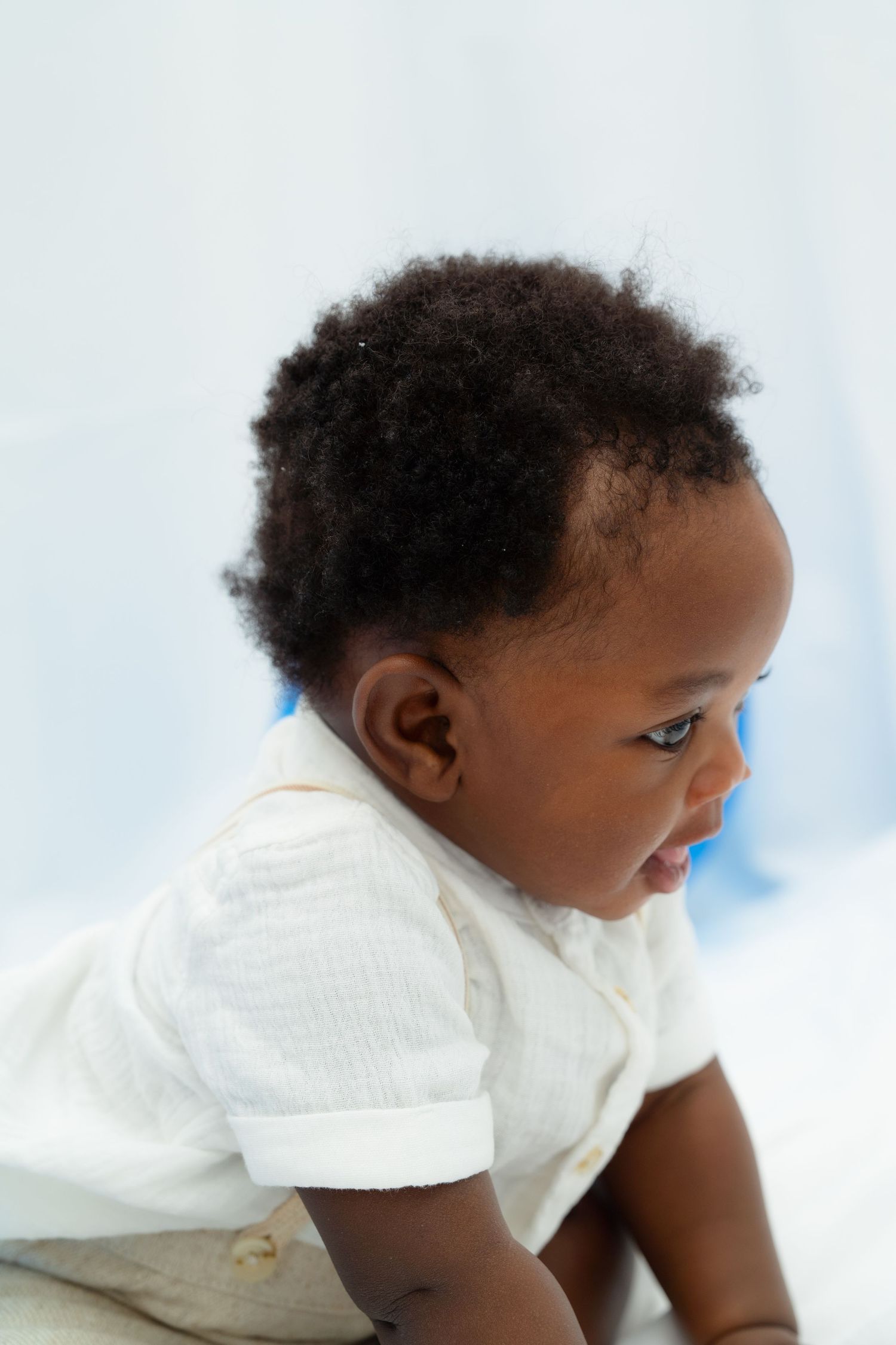 A small child wearing a white shirt looks sideways while crawling against a light background.