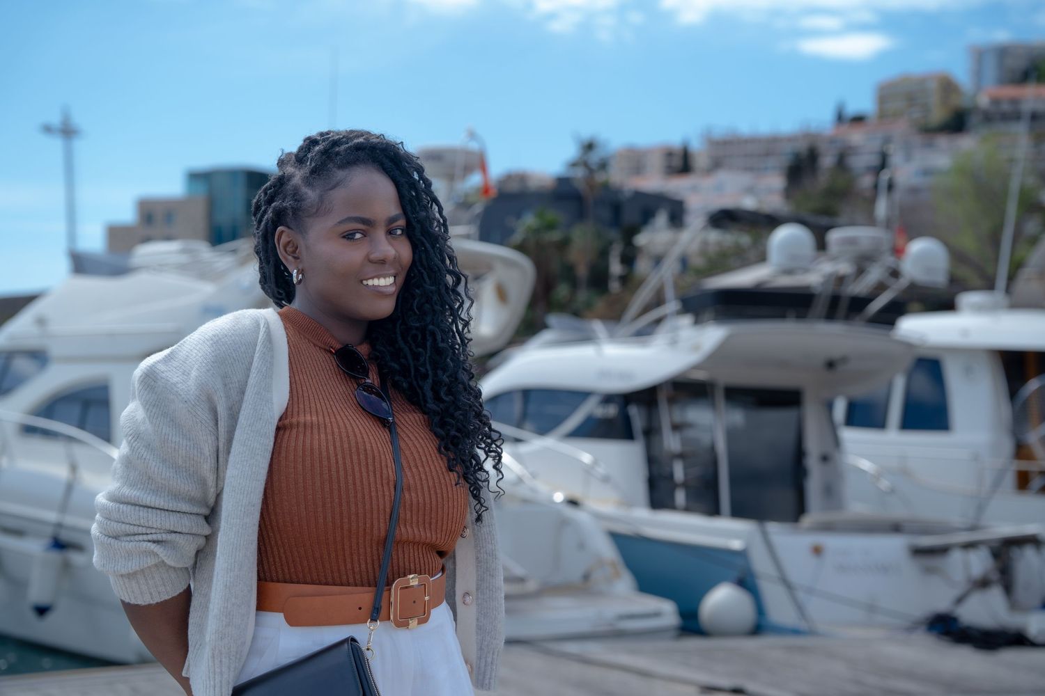 A person in a white cardigan and orange top stands at a marina with boats docked in the background on a sunny day.