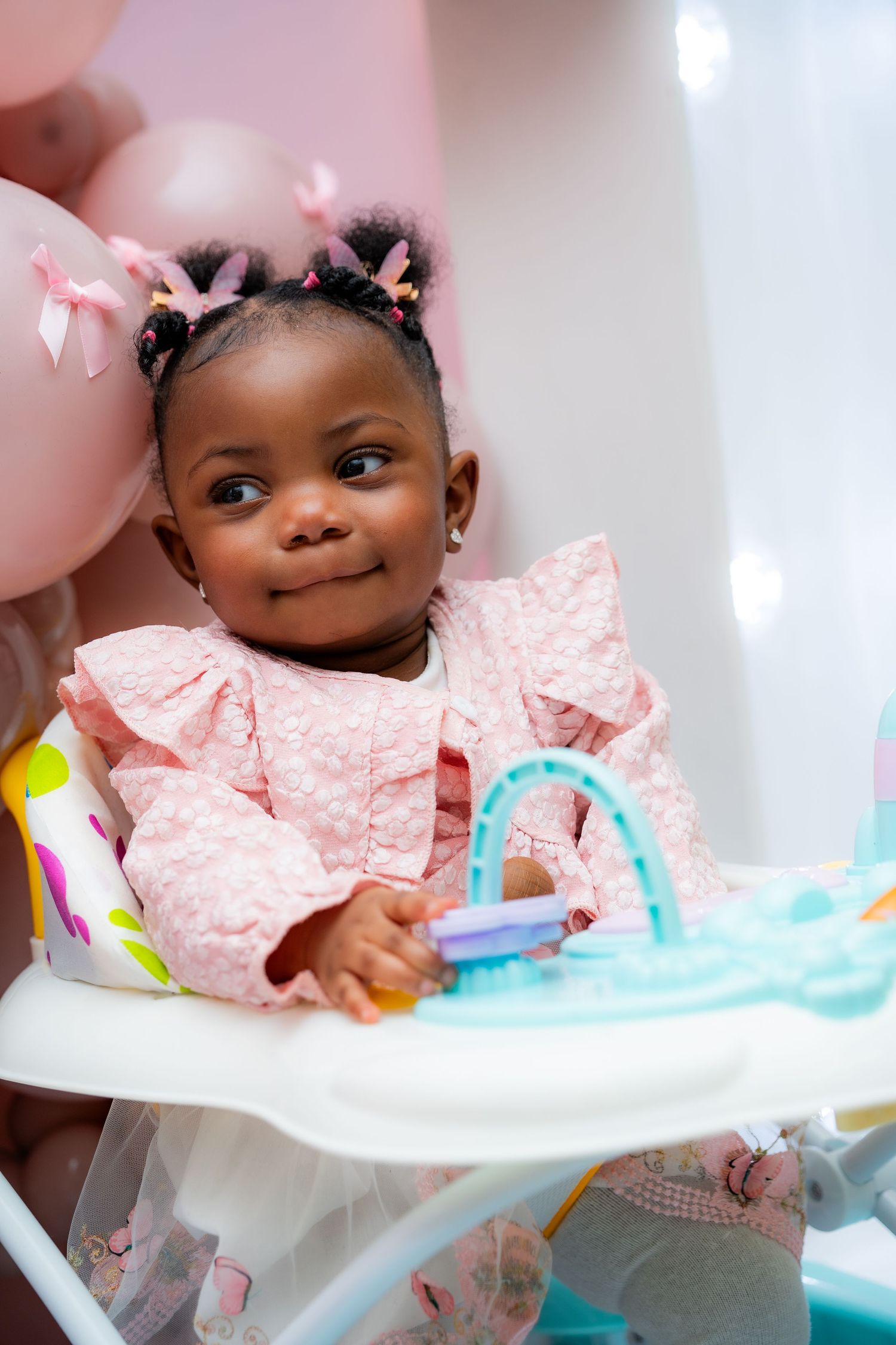 Baby in pink polka dot dress sits in high chair during celebration with pink balloons and decorations.