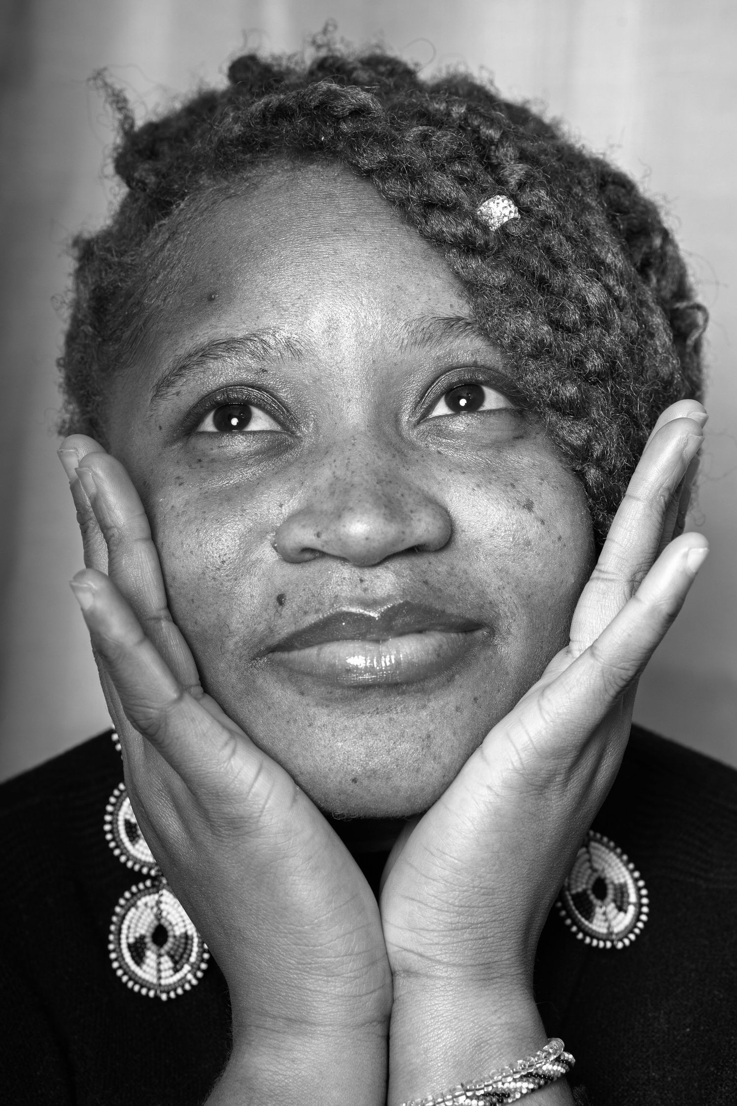 Black and white portrait of someone holding their face with both hands while wearing round dangling earrings and smiling warmly.