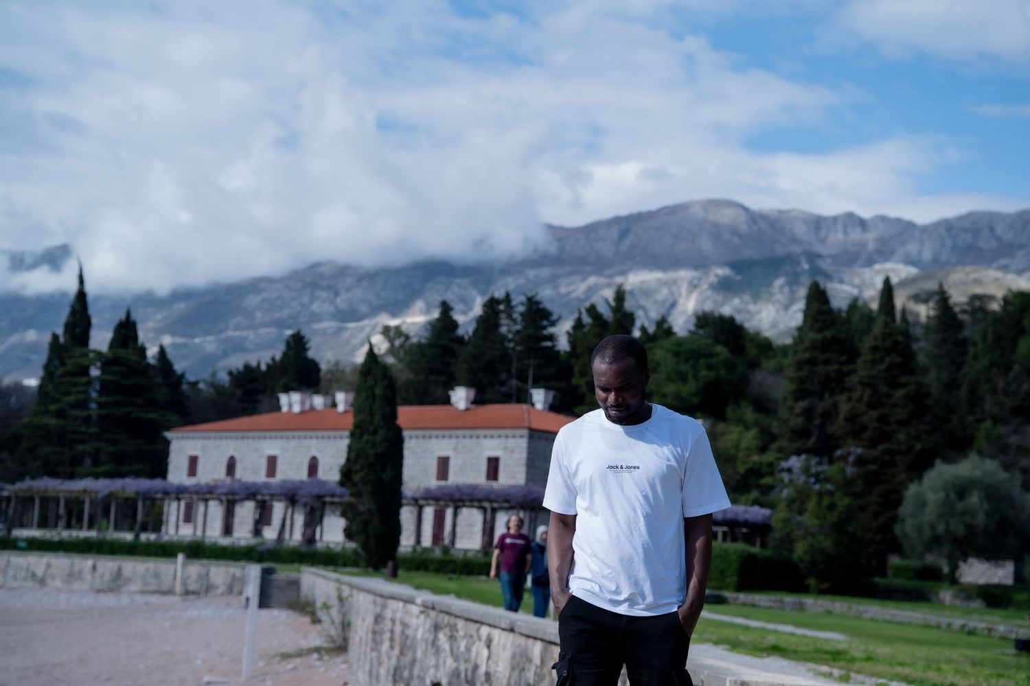 A person stands on a stone wall with cypress trees and Mediterranean villas against a cloudy mountain backdrop.