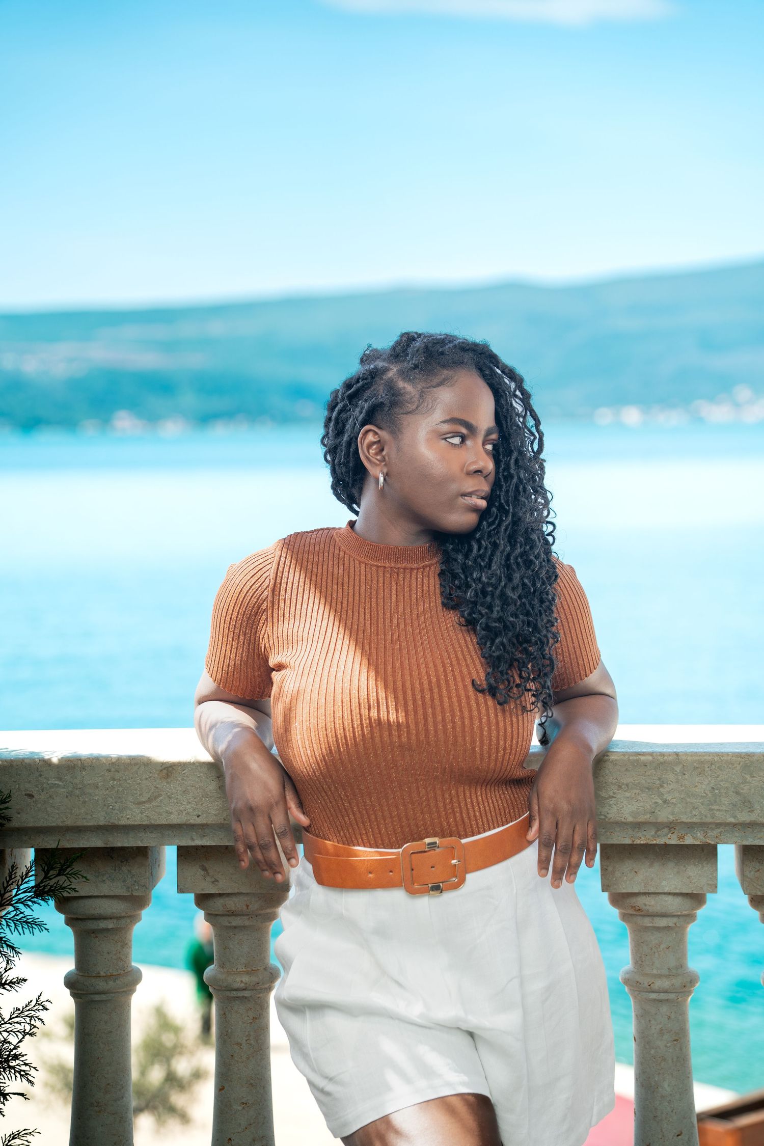 Stylish outfit with orange ribbed sweater and white shorts against scenic coastal backdrop and turquoise waters.