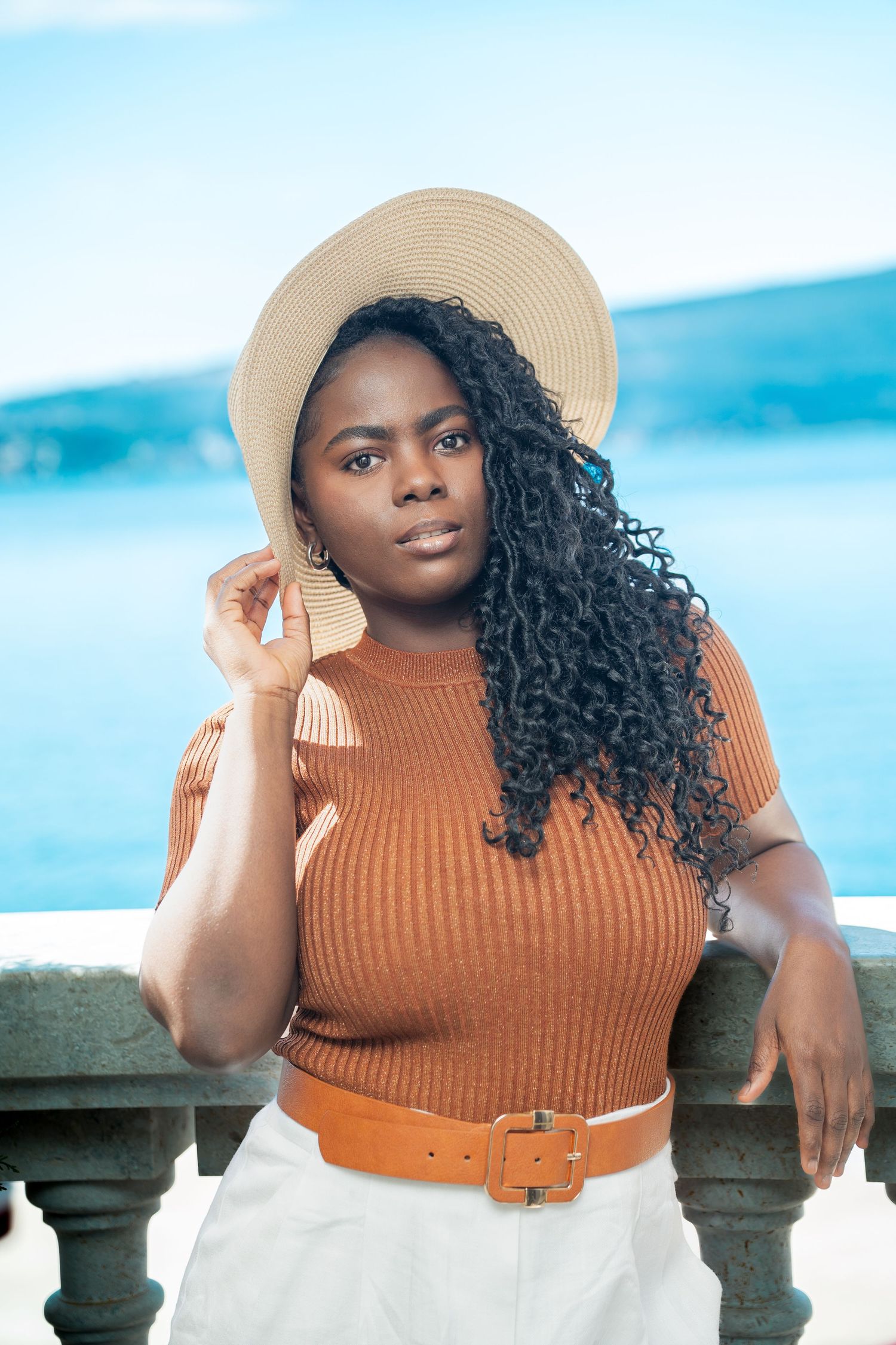 Stylish summer look featuring a tan straw hat, ribbed orange top, and white skirt with brown belt against blue water backdrop.