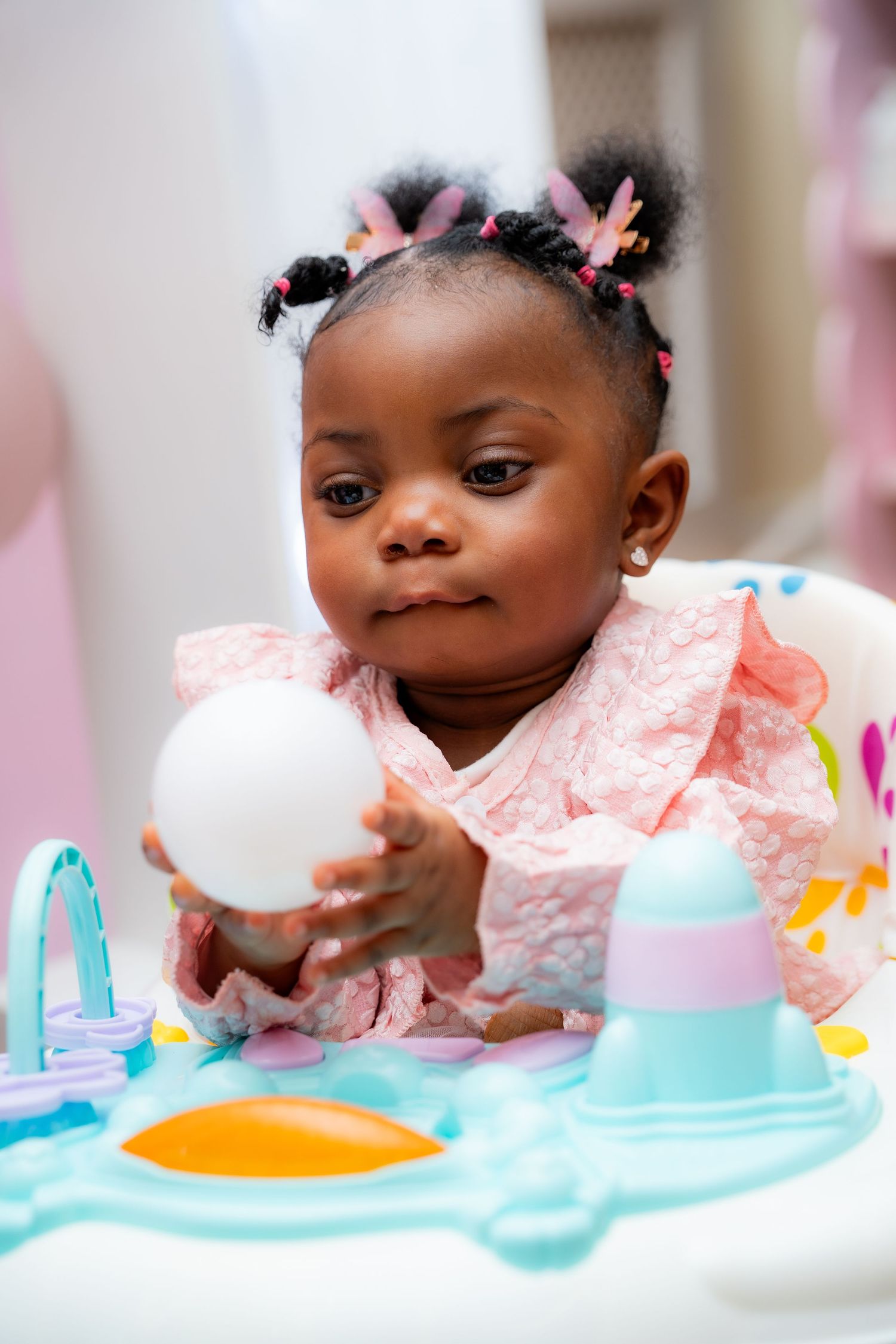Baby in pink dress plays with toys at activity center while wearing cute hair bows in pigtails.