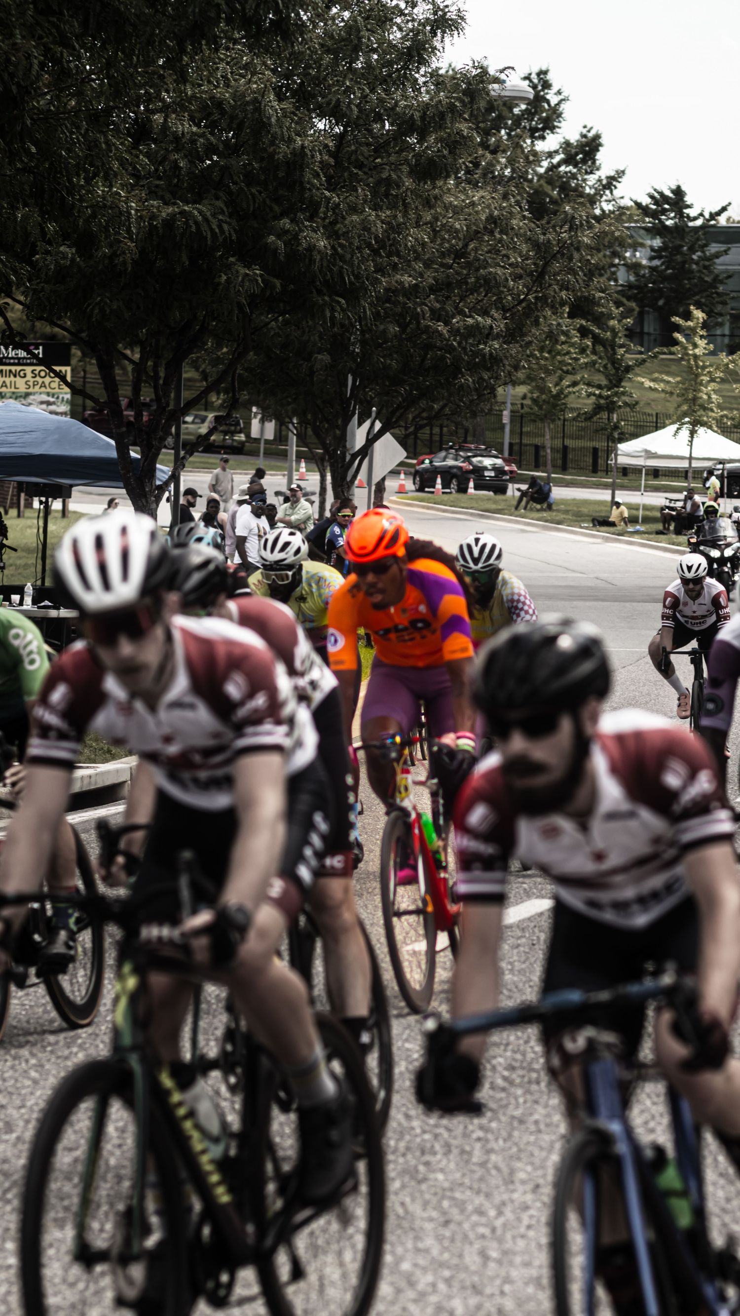 Cyclists in a competitive race wearing helmets and jerseys ride together in a tight group on a road course.