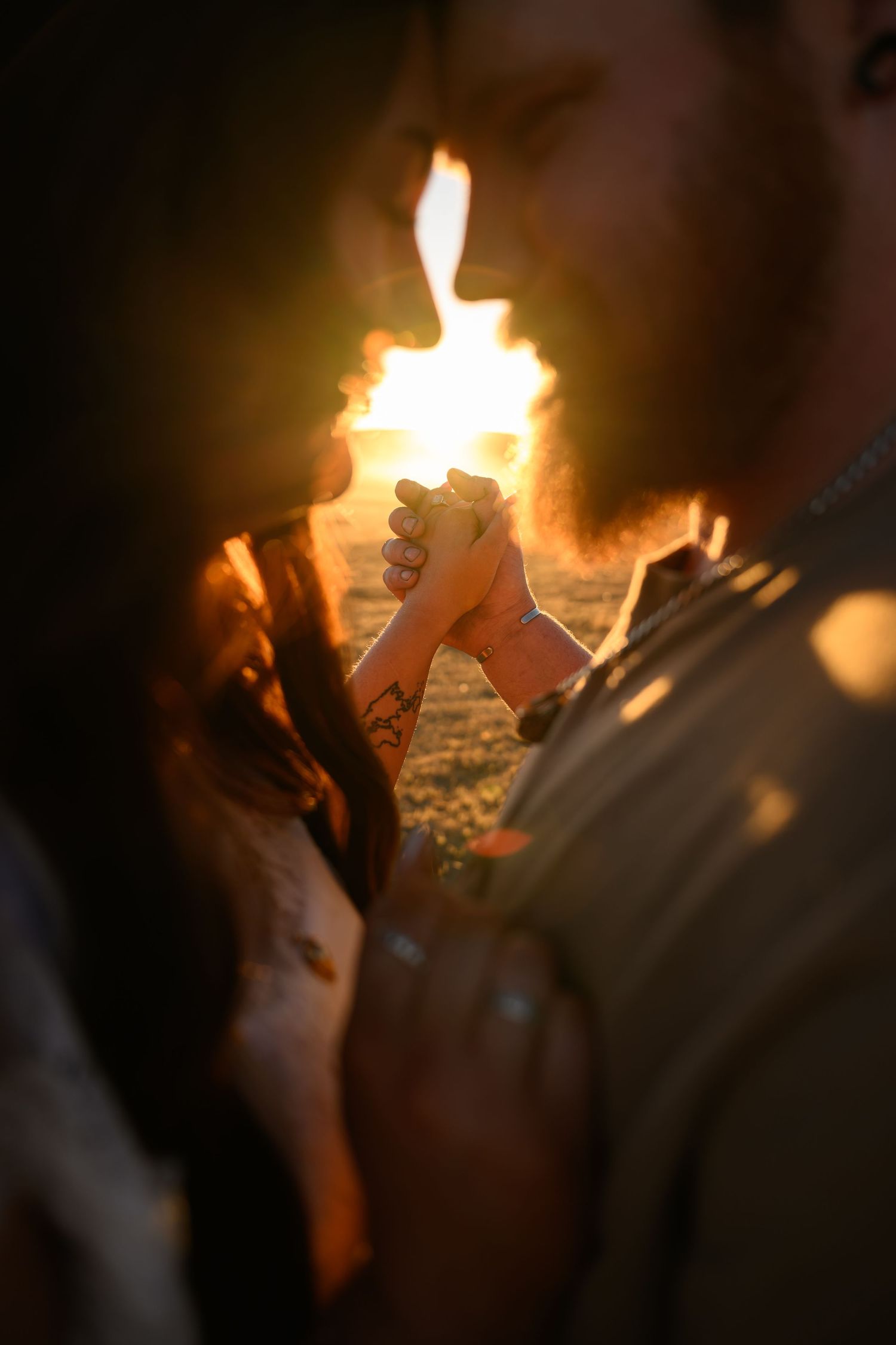 Couple holding hands backlit by golden bonfire flames, with tattoos visible, sharing an intimate romantic moment outdoors.