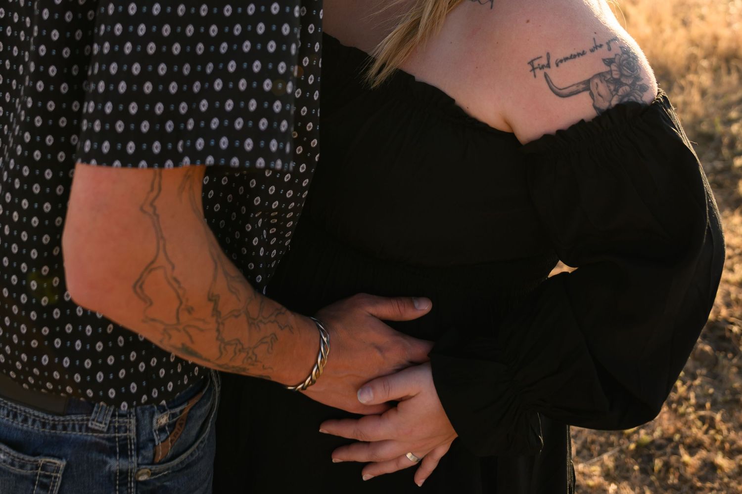 Couple embracing outdoors, both with tattoos, woman in black top, man in patterned shirt and jeans, golden field background.