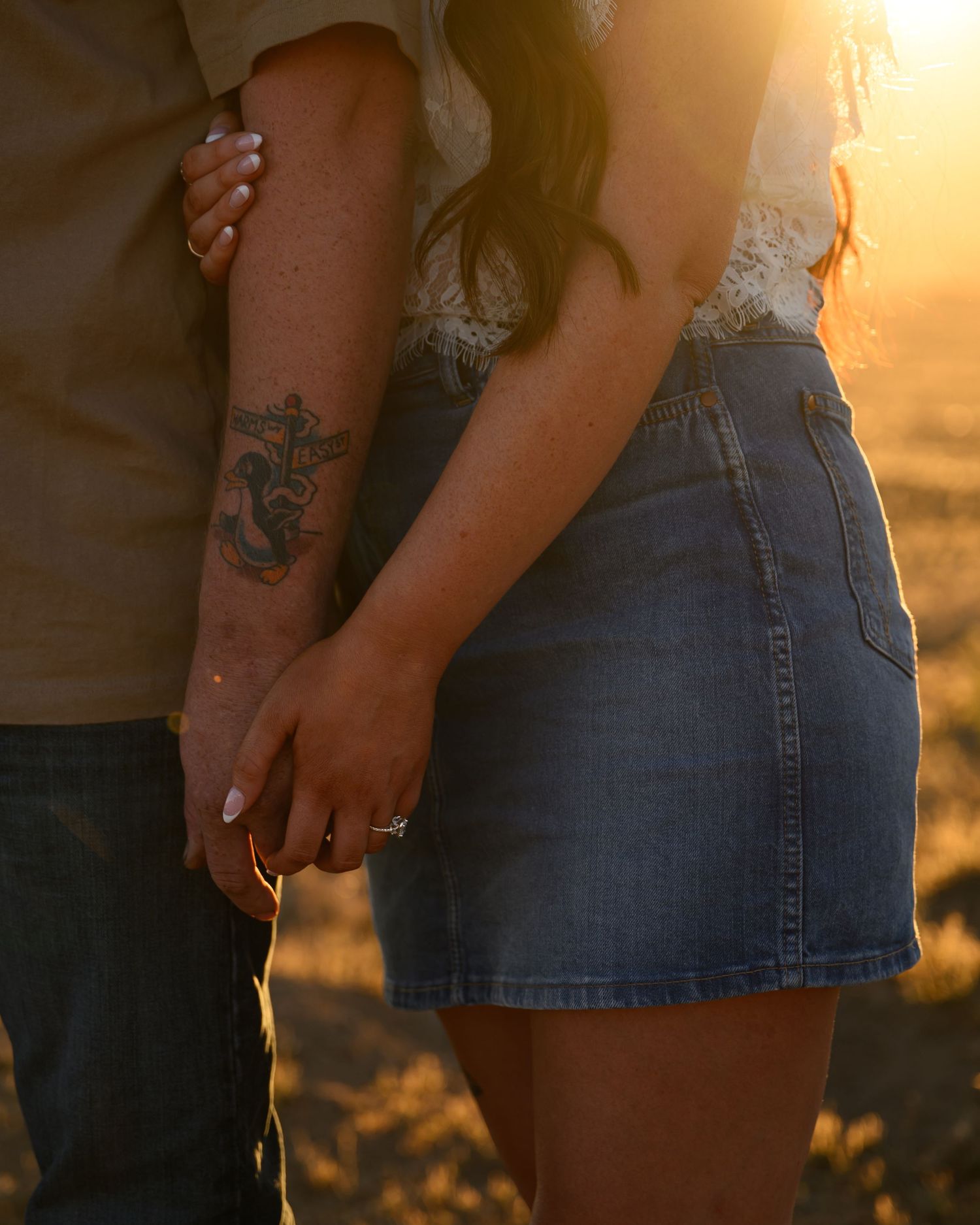 Couple holding hands at golden hour, man with anchor tattoo, woman in denim skirt, romantic outdoor engagement photo.