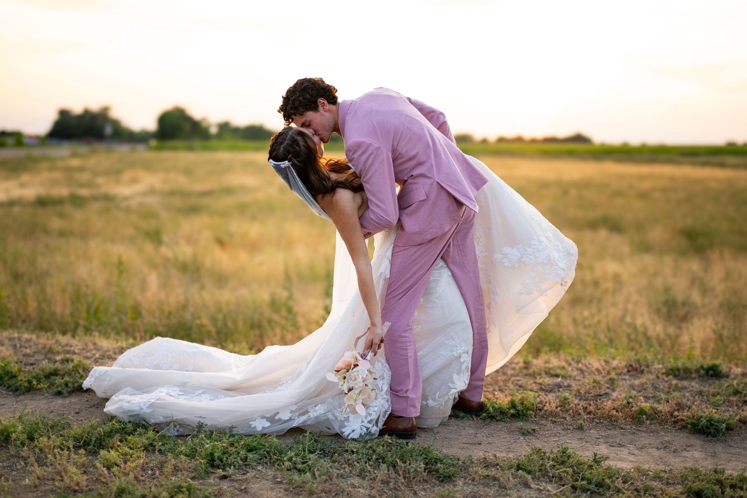 A couple in wedding attire share a romantic moment in a golden field at sunset.