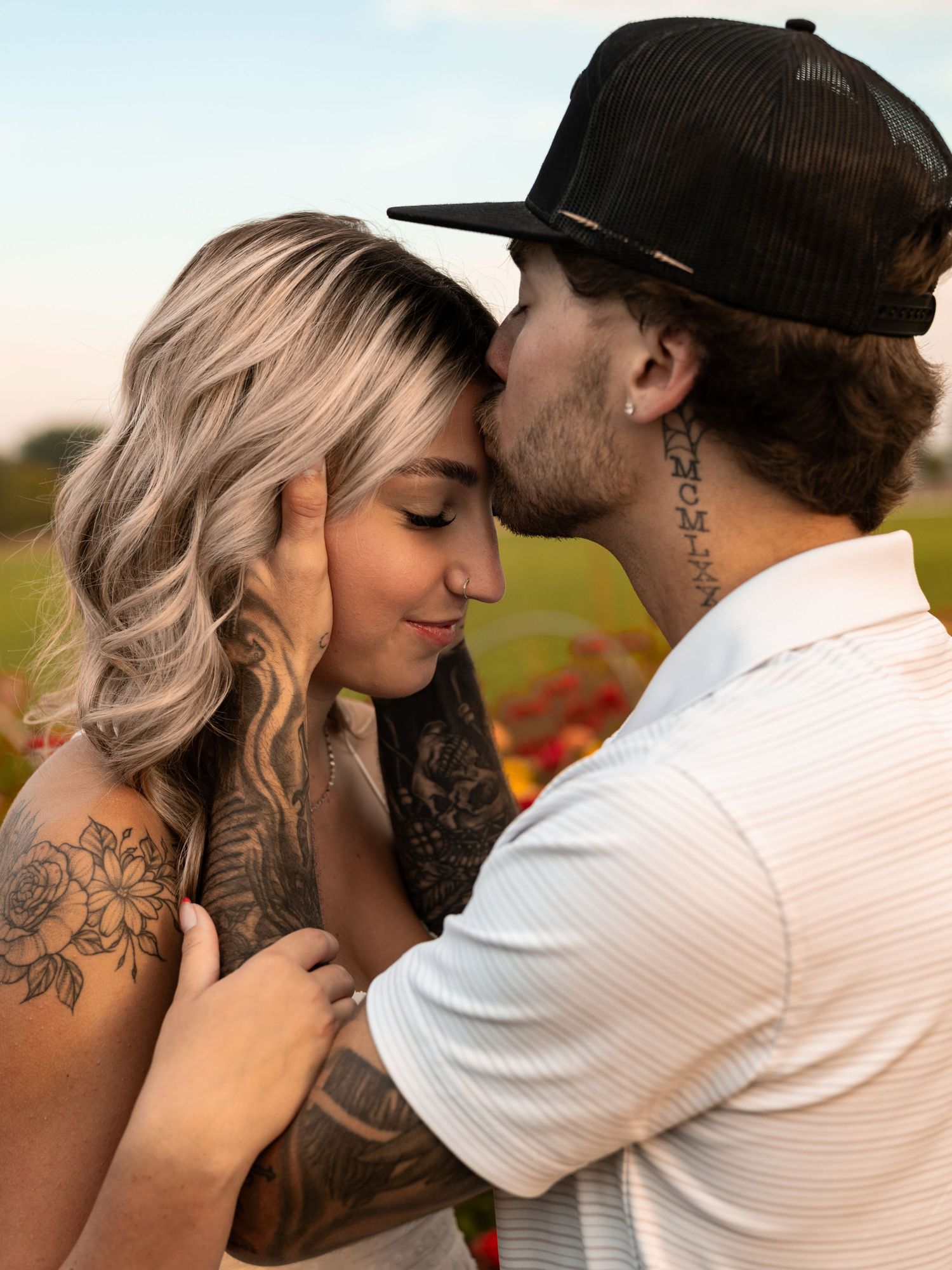 Romantic couple embracing in a field during sunset with visible tattoos and casual attire.