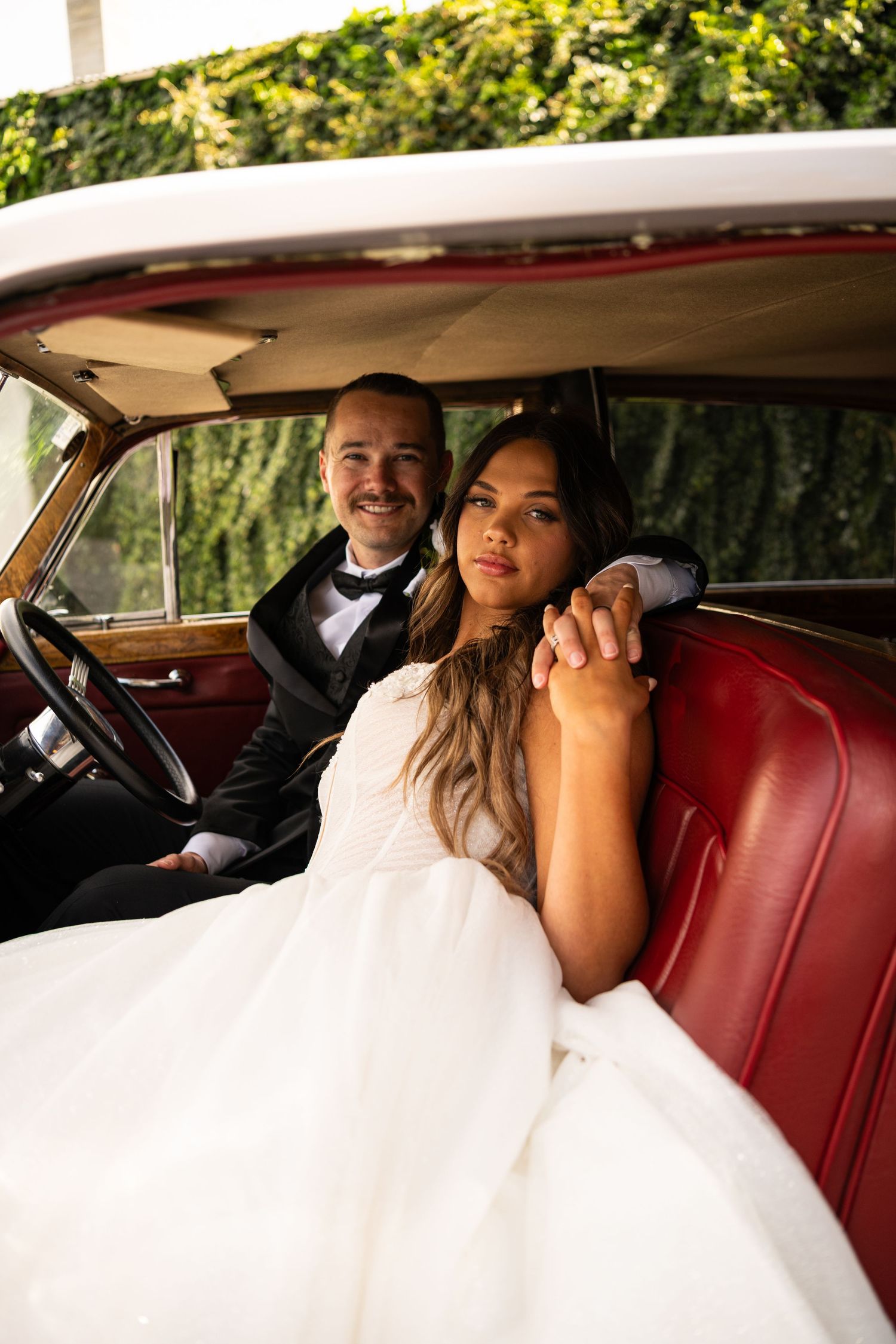 A romantic wedding couple shares intimate moments in the back seat of a vintage car with red leather interior.