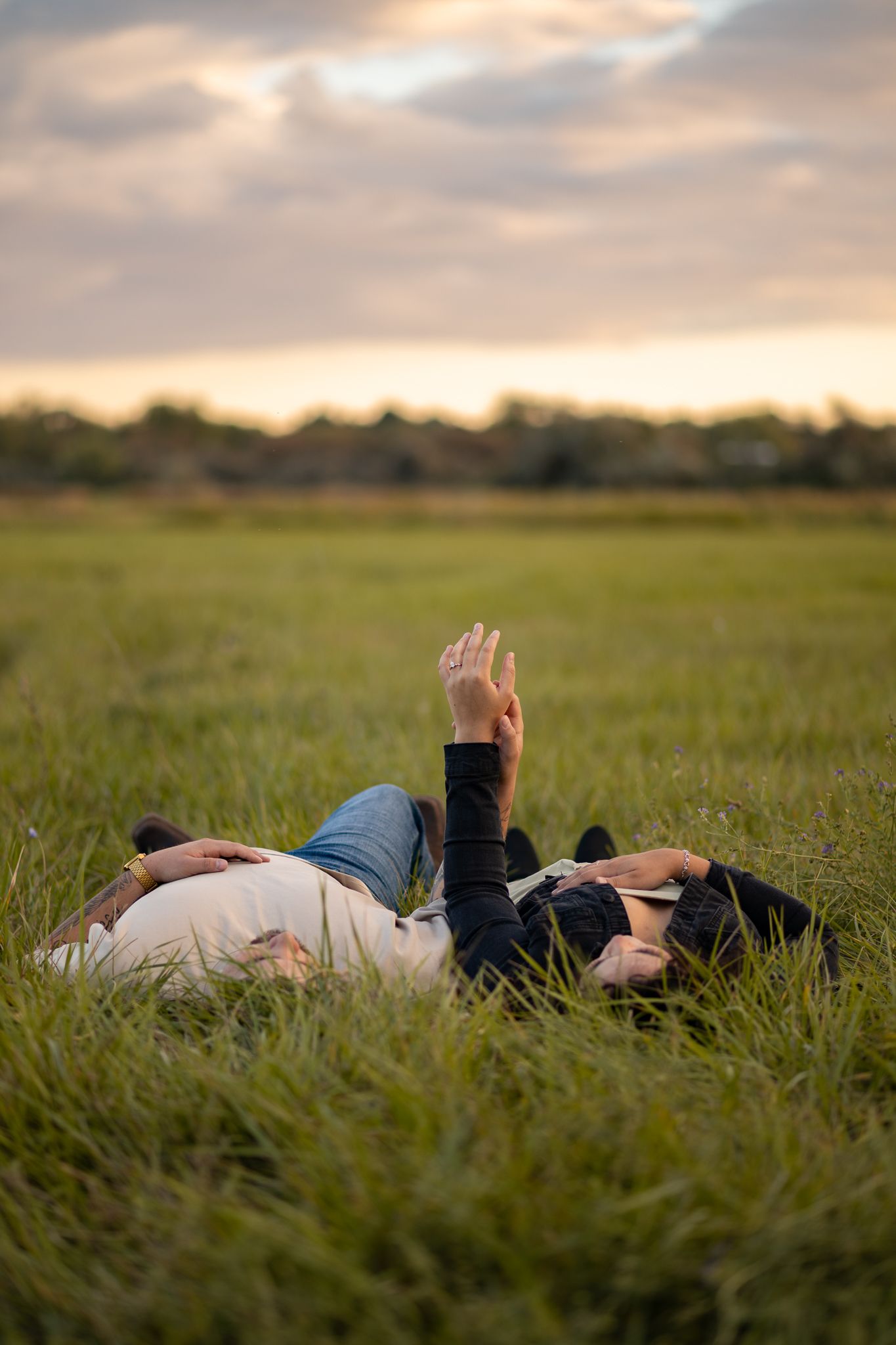Two people lie peacefully in a lush green field at sunset, surrounded by tall grass swaying in the breeze.