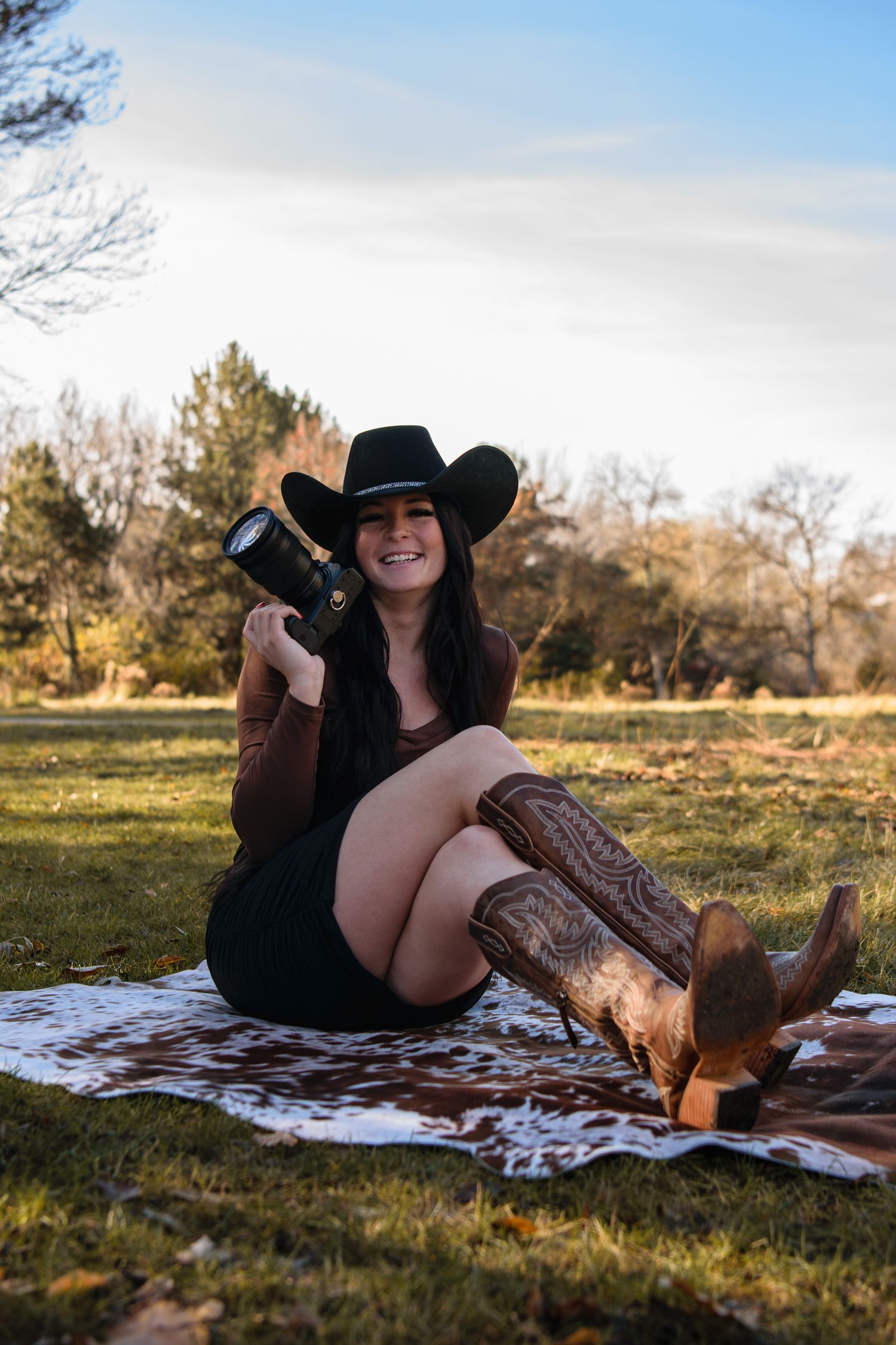 A photographer in cowboy boots and black hat sits on a blanket outdoors holding a professional camera.