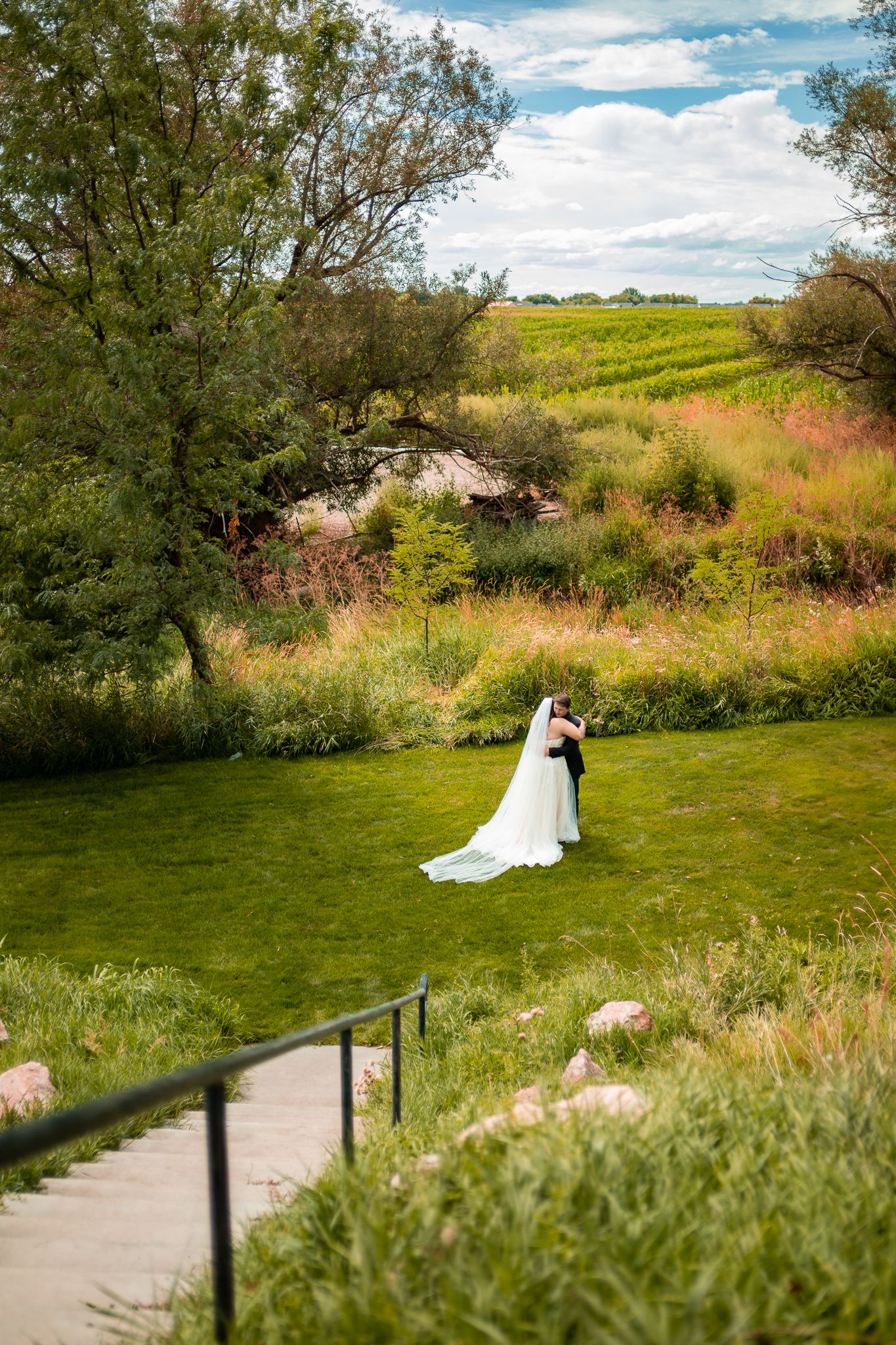 A couple in wedding attire embraces on a grassy hillside path surrounded by natural vegetation on a sunny day.
