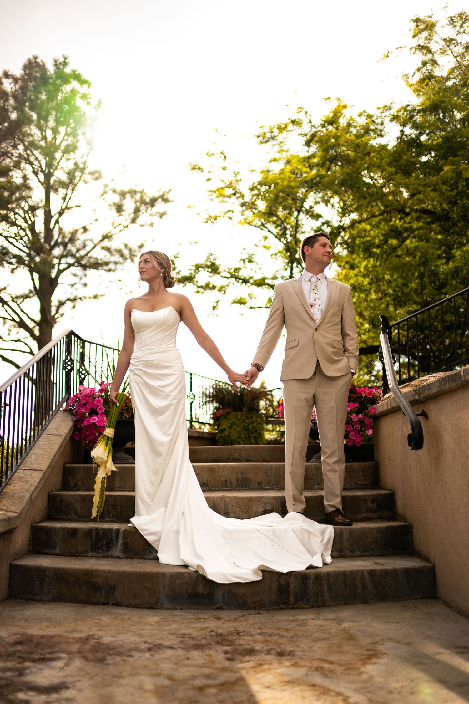 A couple in wedding attire holding hands while standing on stone steps with pink flowers and greenery in the background.