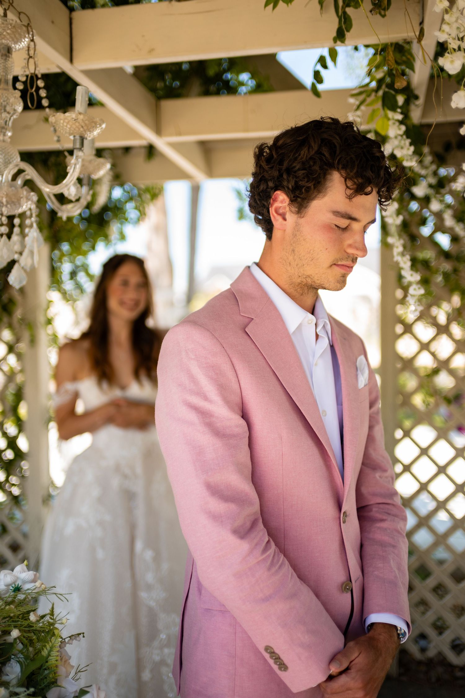 A person in a pink suit jacket and white shirt stands in a garden setting with white lattice and greenery in the background.