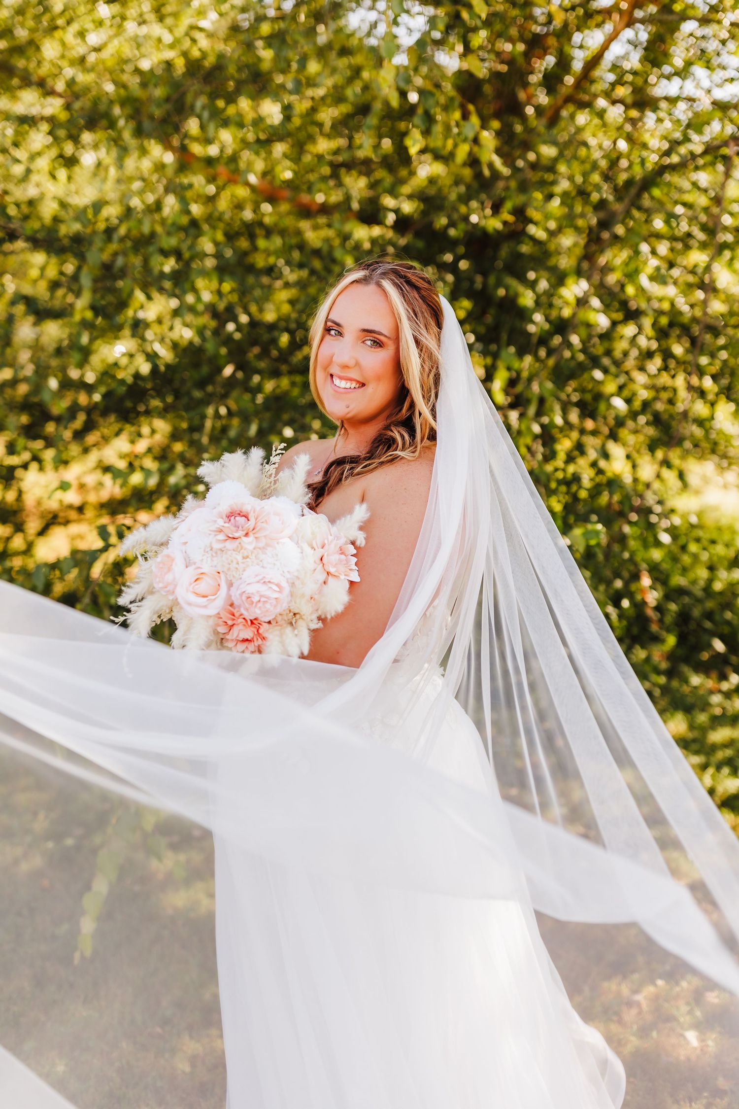 Bride in flowing white veil poses with pink rose bouquet against sunlit trees.