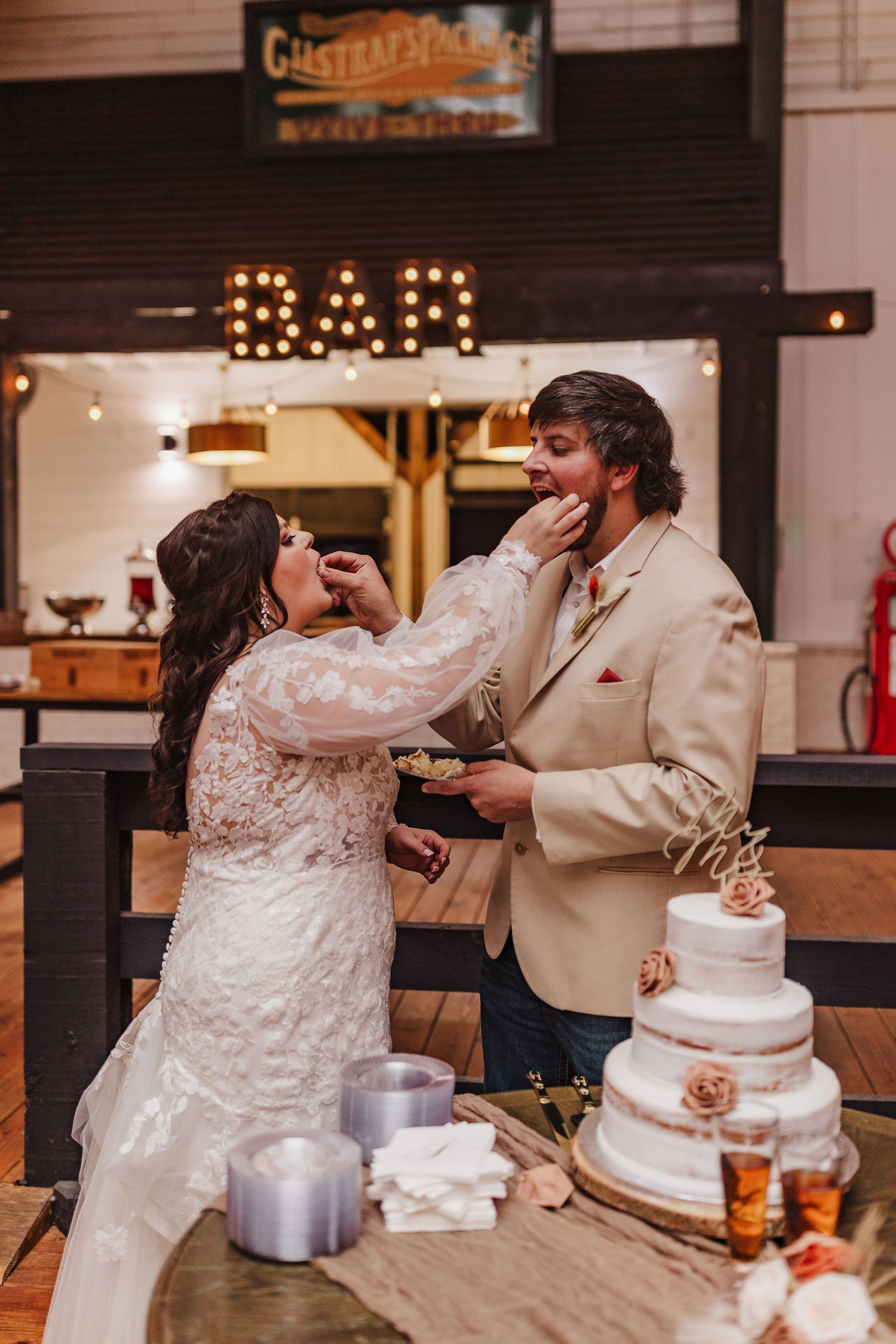 Wedding cake cutting ceremony at LL Farm Events Venue with rustic decor and an illuminated bar sign.