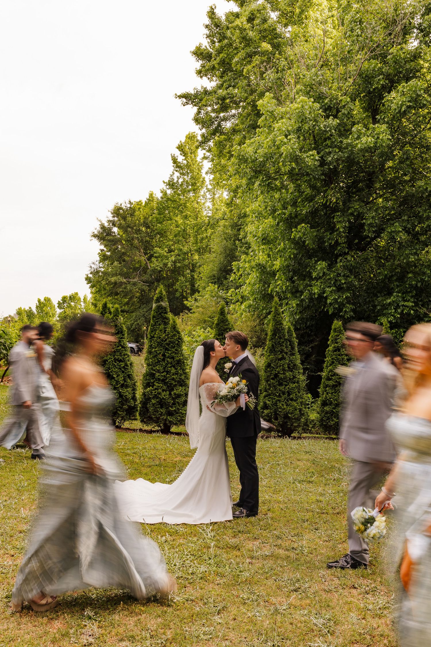 Bride and groom kissing outdoors surrounded by blurred wedding party in silver attire with lush green trees backdrop.