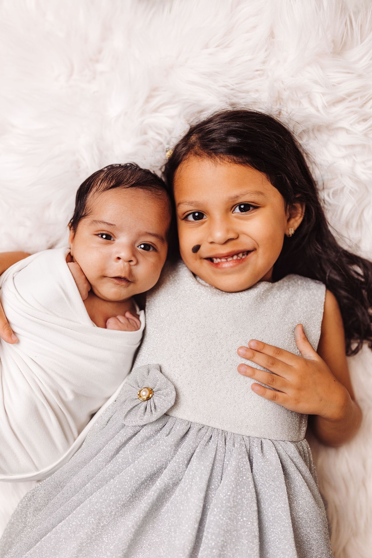 Two children laying on white bedding wearing matching gray and white dresses share sweet smiles together.