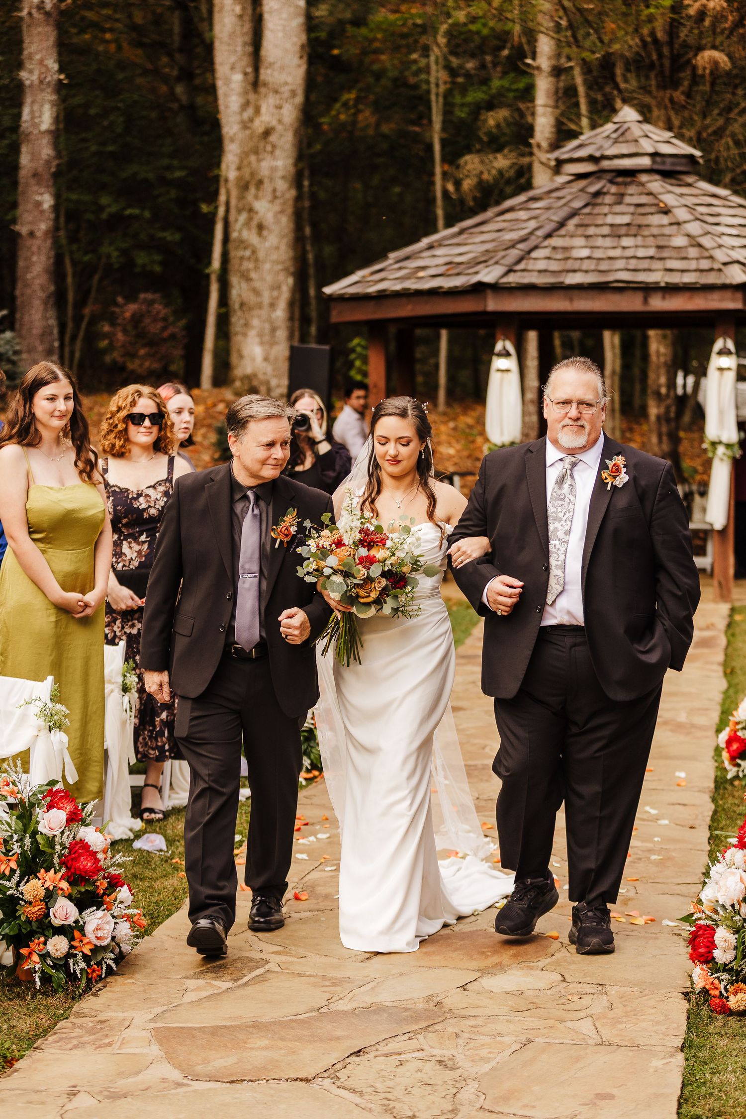 Wedding procession moves along a rustic stone pathway surrounded by autumn trees and floral decorations.