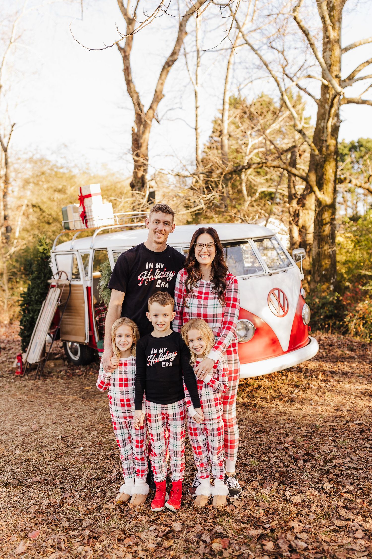 Family of five in matching red plaid Christmas pajamas posing in front of a vintage VW bus with holiday gifts on top.