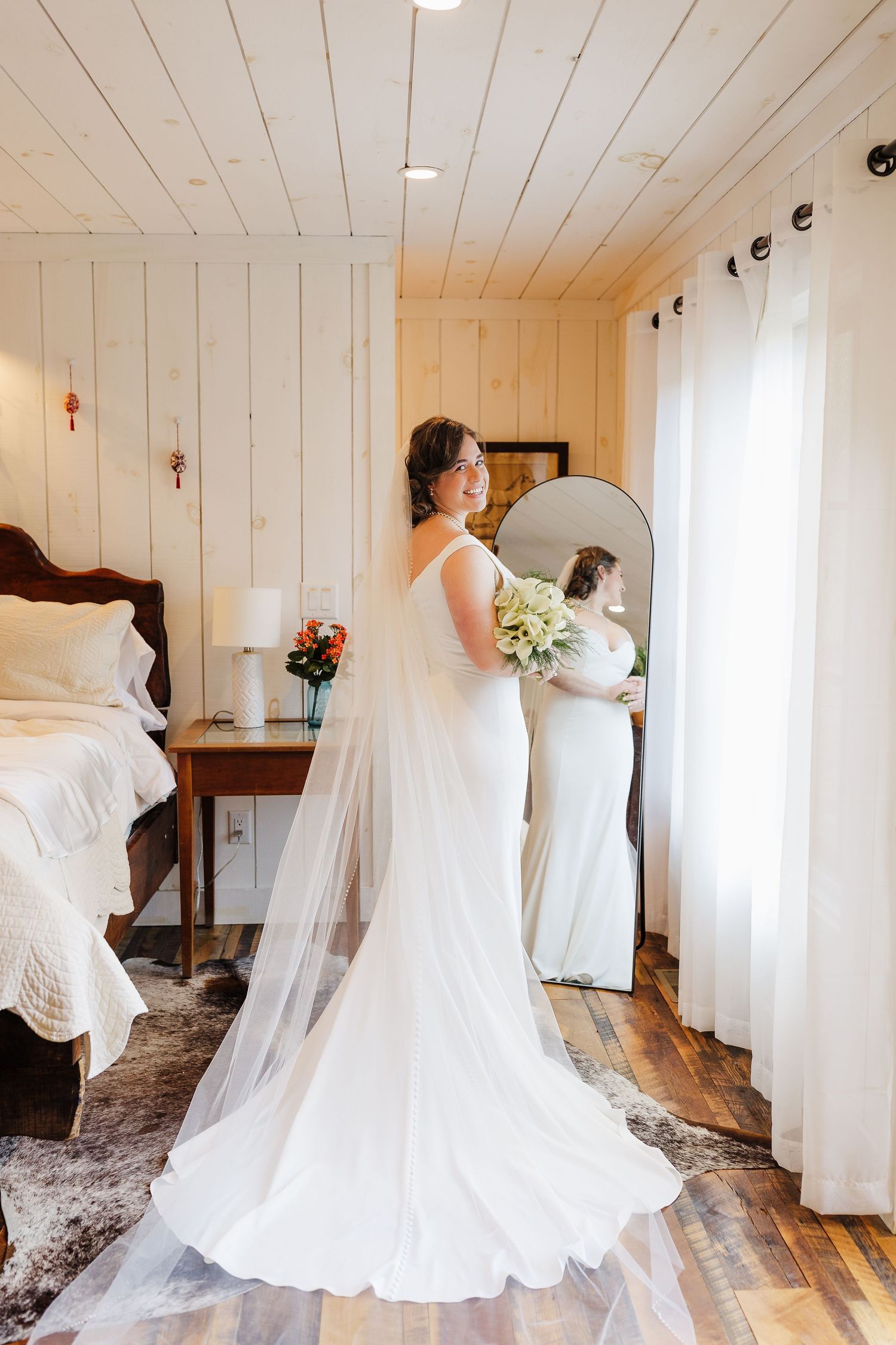 Bride in white gown and veil holding bouquet, smiling while viewing her reflection in a round mirror in a rustic room.