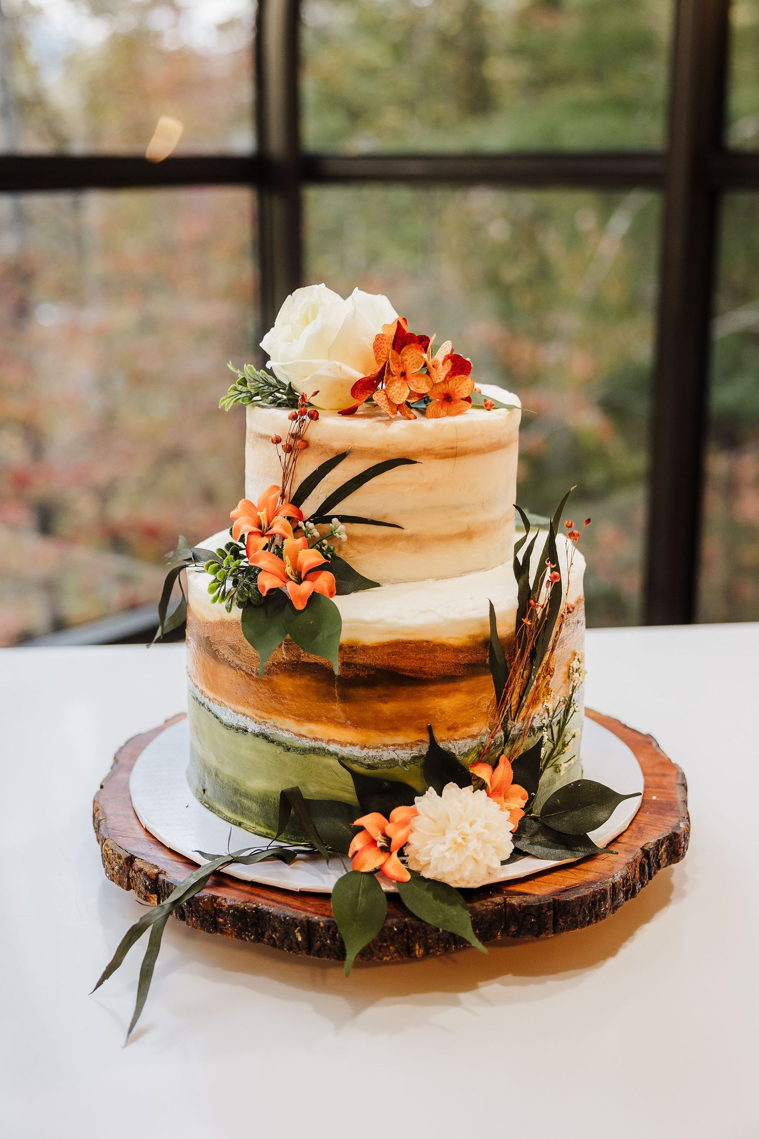 A rustic wedding cake with peach flowers sits on a wood slice base against a window backdrop.