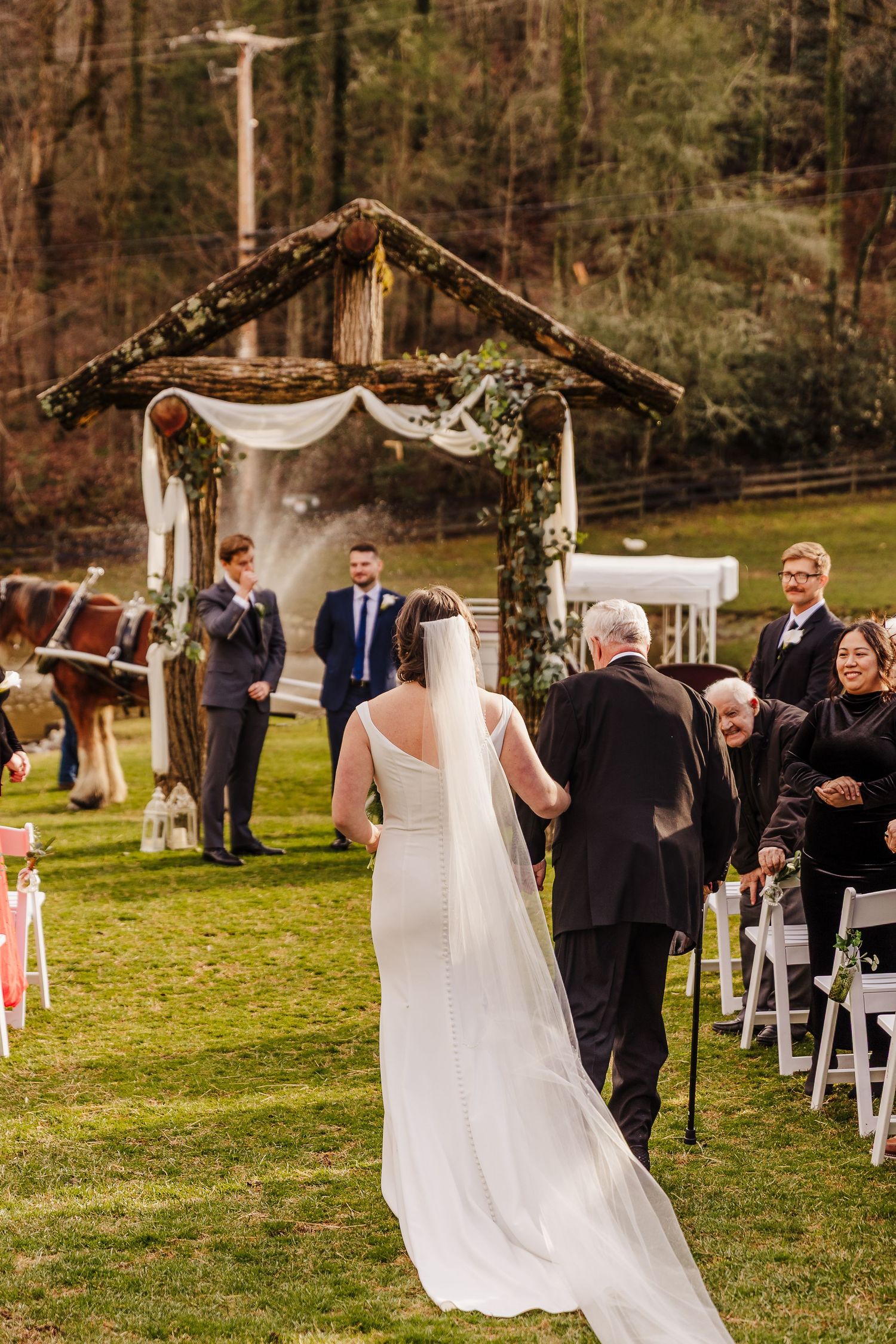 Bride walks down outdoor aisle with father toward rustic wooden arch at elegant garden wedding ceremony.