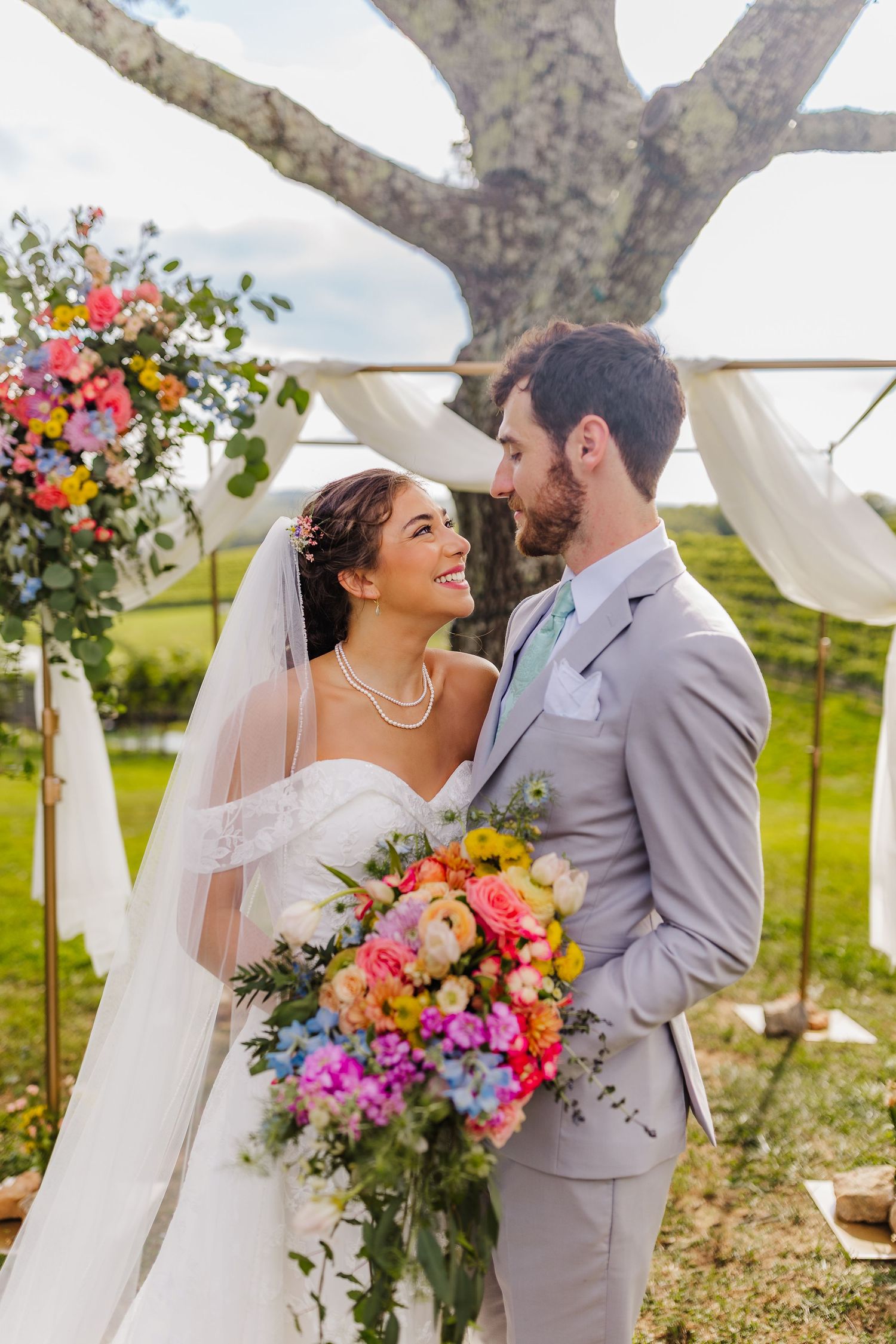 Bride and groom share a romantic moment at outdoor wedding ceremony with colorful floral arch and lush green backdrop.