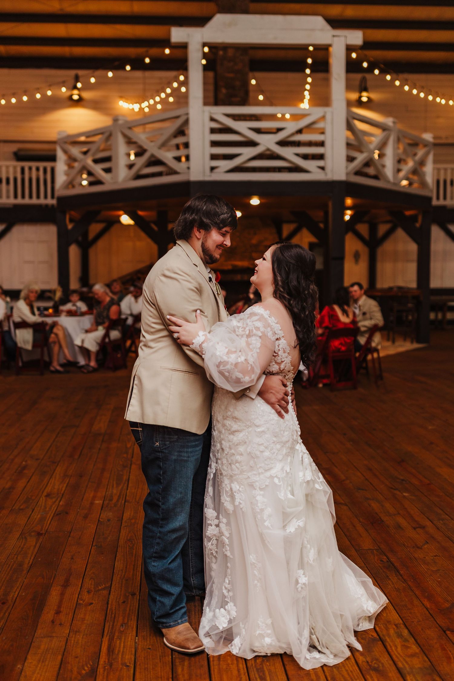 Bride and groom share their first dance on a wooden floor in a rustic wedding venue with string lights and guests seated nearby.
