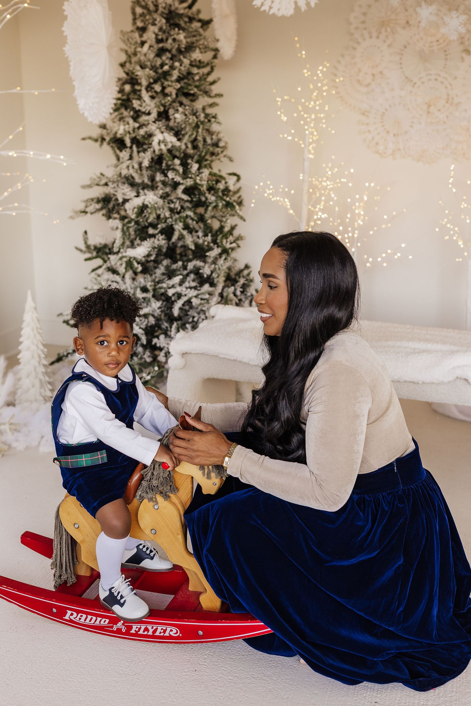 A mother helps her toddler sit on a Radio Flyer rocking horse near a decorated Christmas tree.