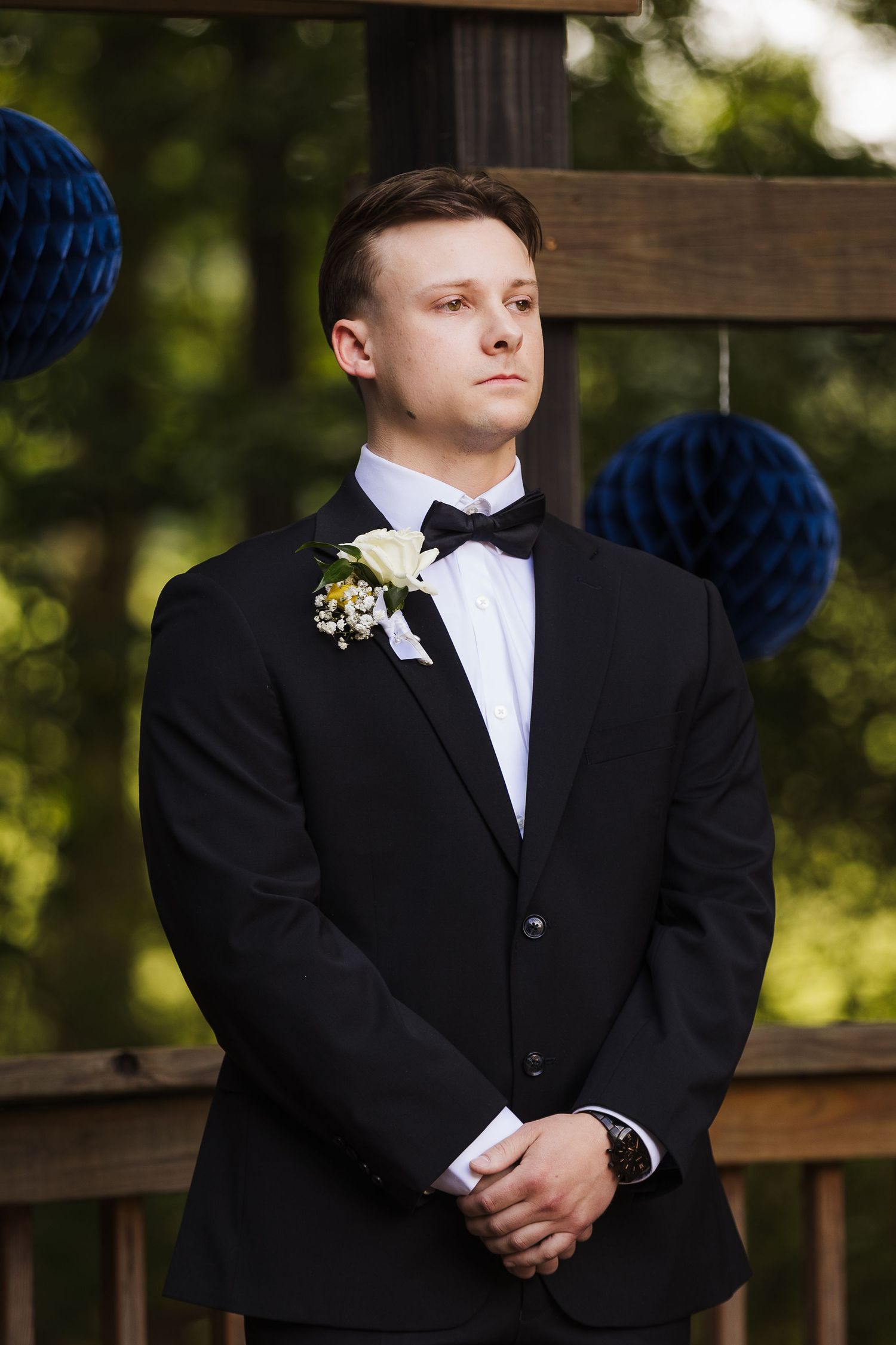 Young groom in black tuxedo with white boutonniere and bow tie stands at outdoor wedding ceremony with blue decorations.