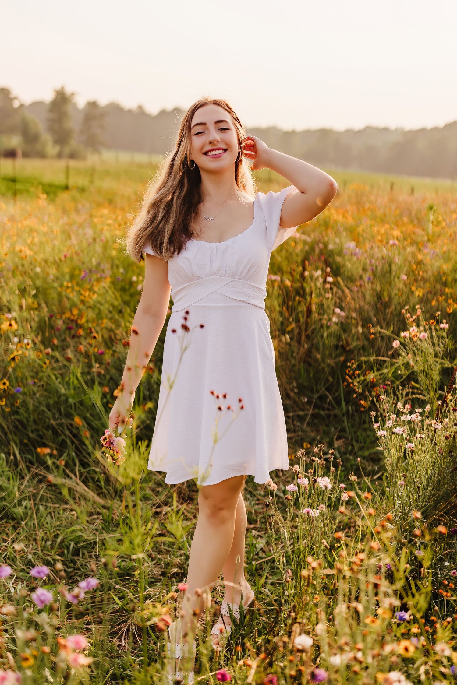 Young woman in white dress smiling in a wildflower meadow during golden hour sunset.