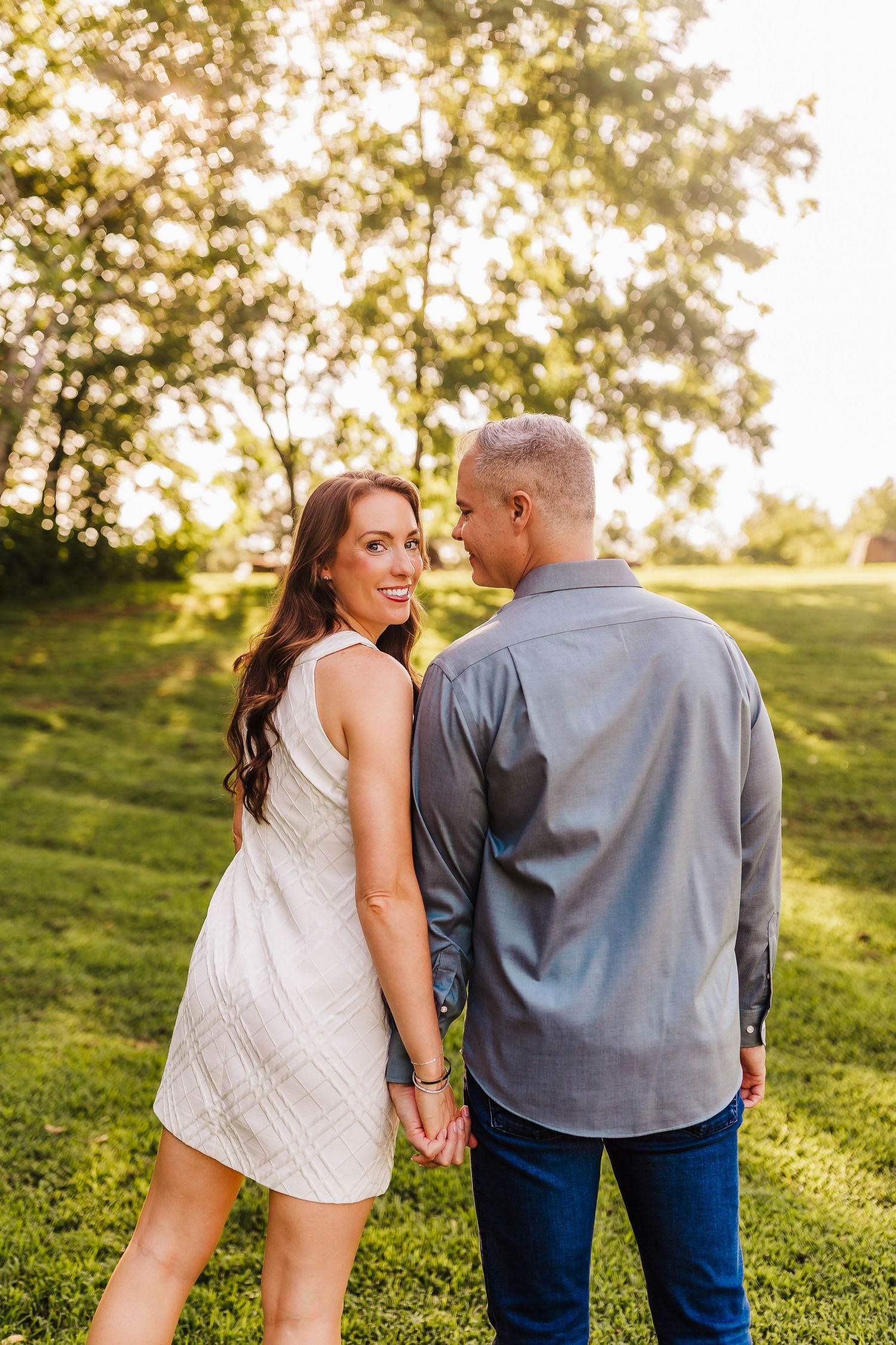 Couple holds hands while walking through sunlit grassy hill during outdoor photo session.