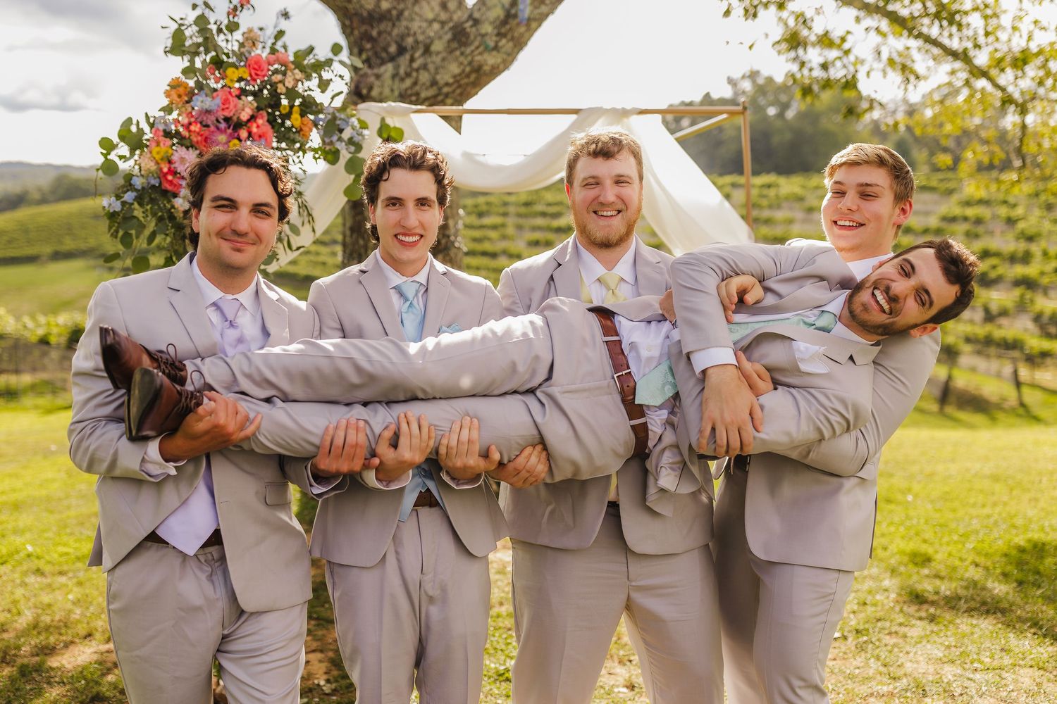 Groomsmen in gray suits carrying the groom outdoors at a rustic vineyard wedding ceremony.
